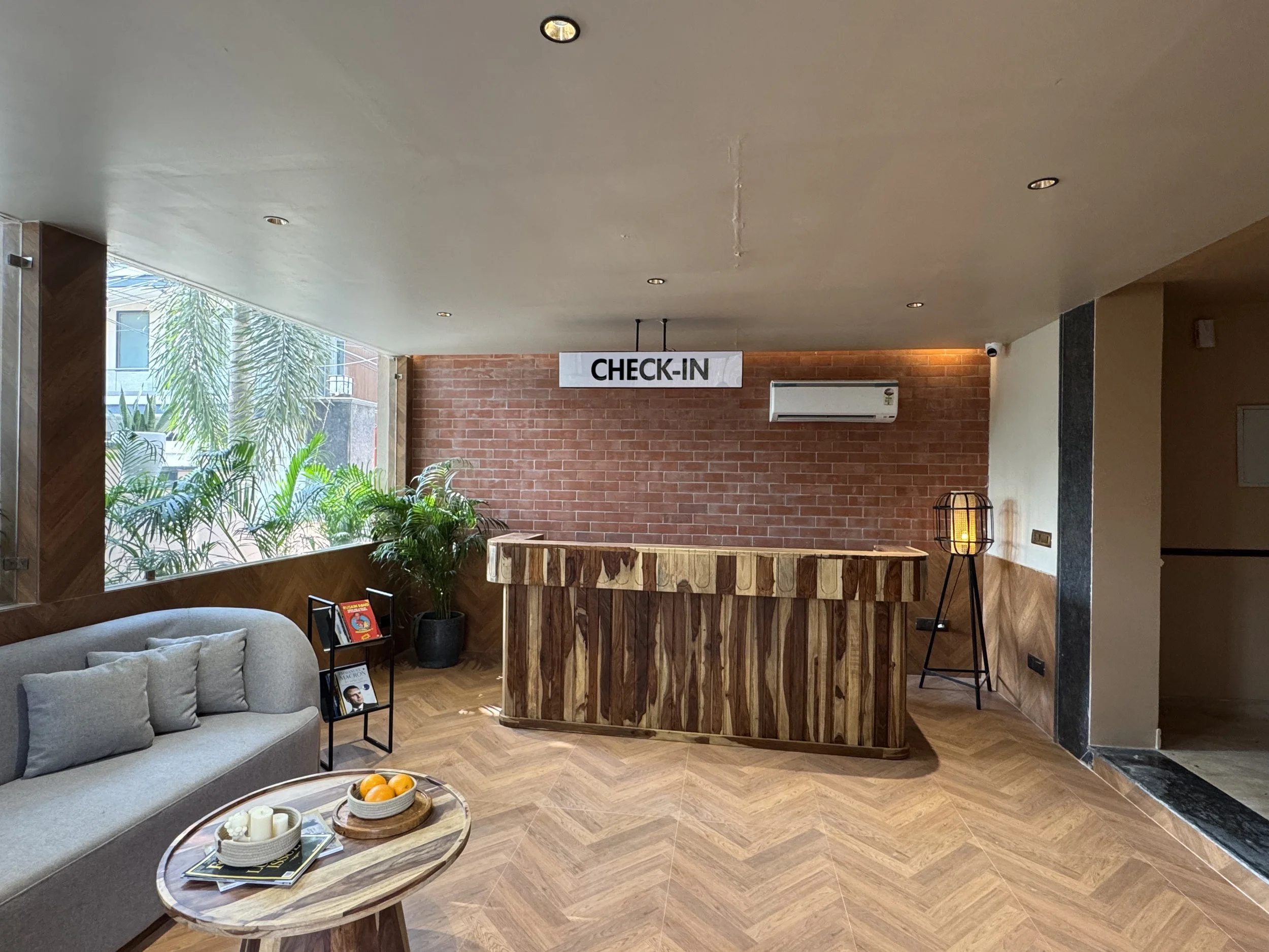 Hotel check-in counter with a sign, a potted plant, a small seating area with a sofa and magazines, in front a round wooden table with candles and oranges.