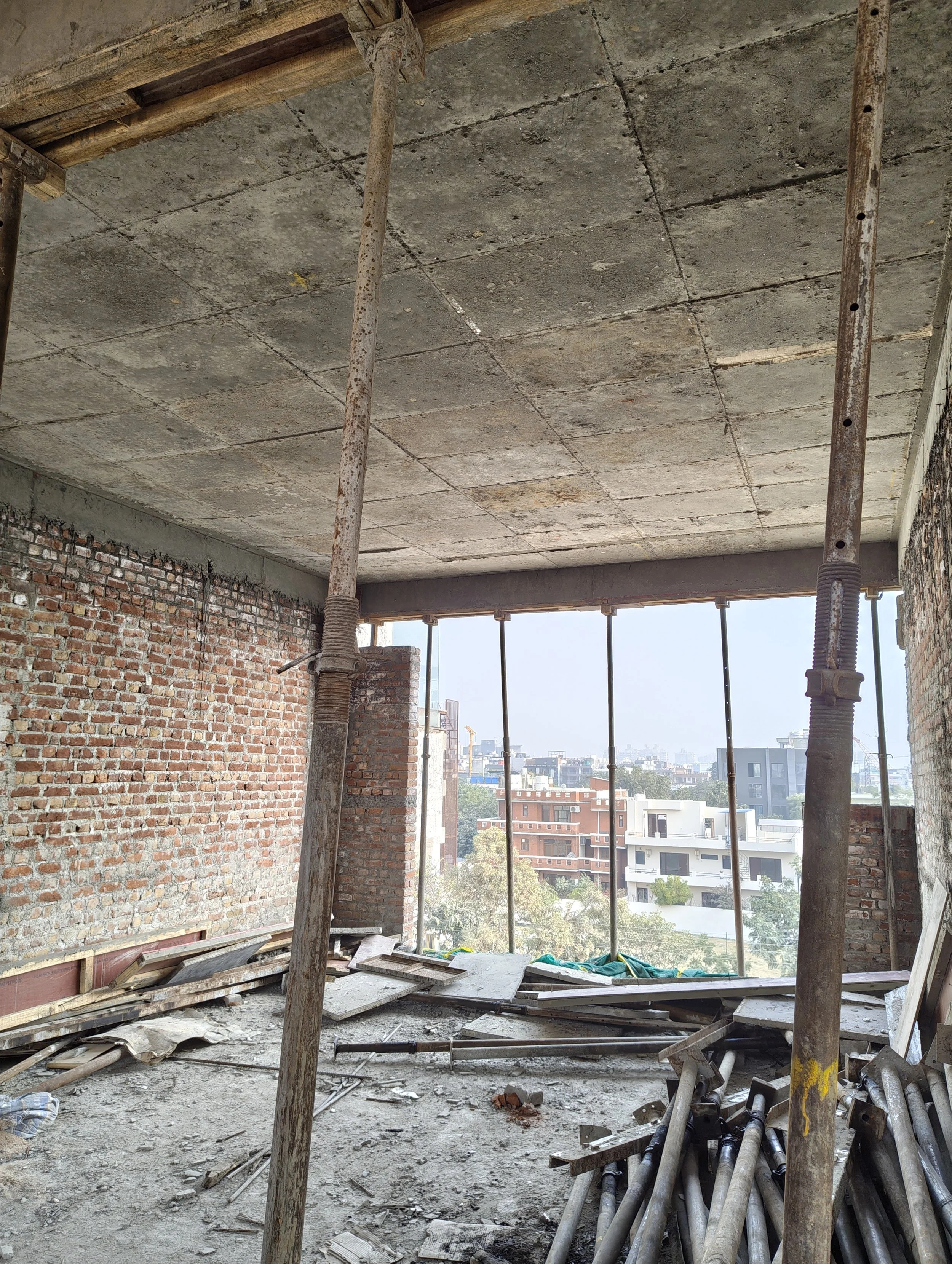 Construction site of a building with brick walls, a concrete ceiling, and metal supporting poles, with cityscape view in the background.