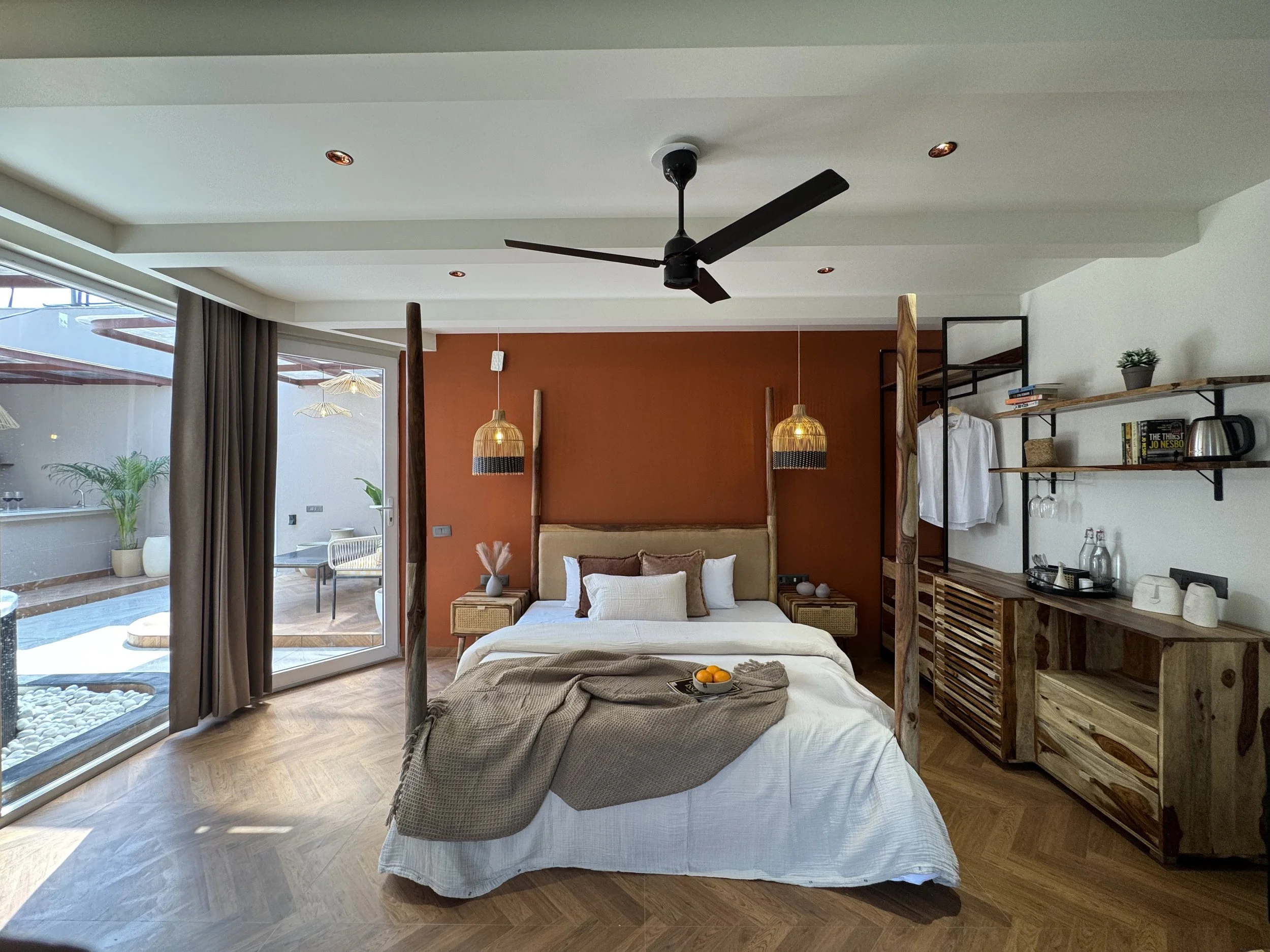 Modern bedroom with a large bed, pendant lights, sliding glass door leading to balcony with outdoor furniture, wooden flooring, and a built-in wooden cabinet with shelves.