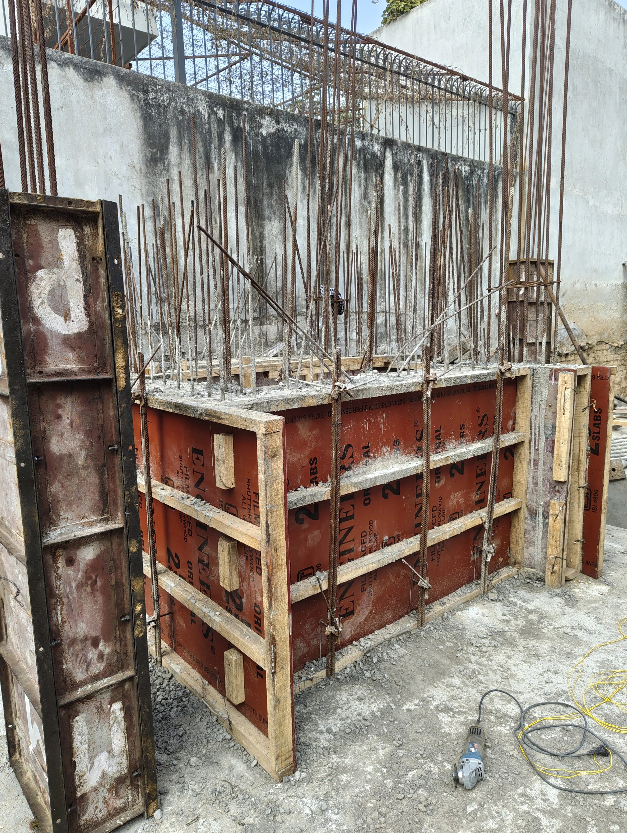 Construction site with reinforced concrete formwork and steel rebar on a dirt ground, with construction tools and wiring nearby.