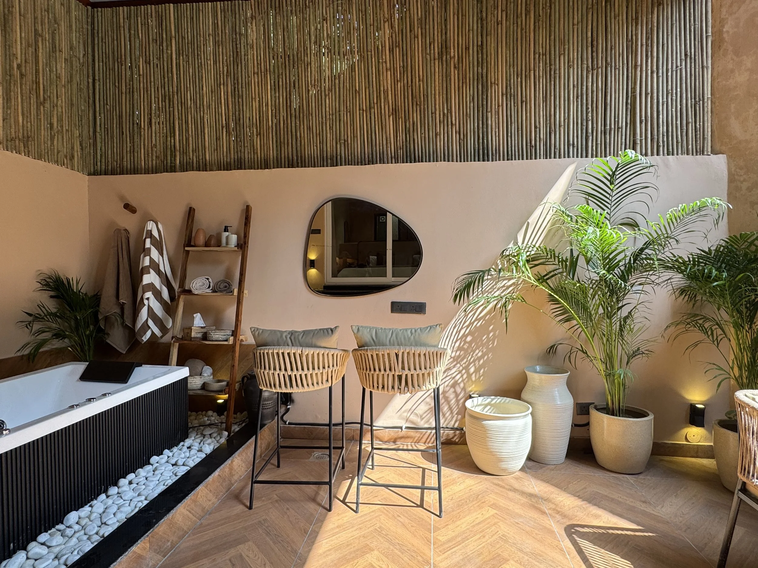 Interior of a spa or wellness room with a large bathtub filled with white stones, wooden floors, potted plants, a ladder shelf with towels and spa products, wicker bar stools with cushions, and a decorative mirror.