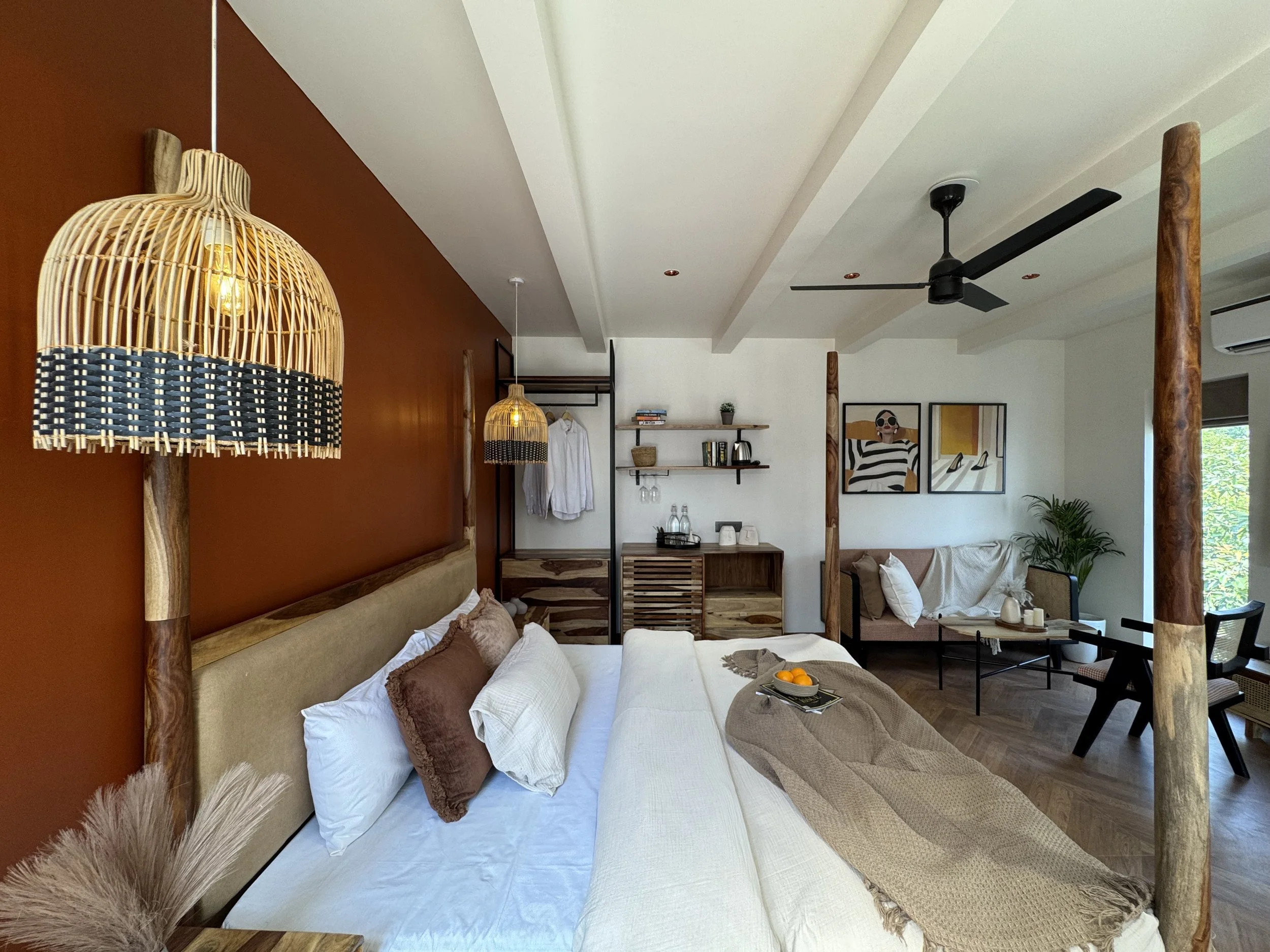 Spacious bedroom with a large bed that has multiple pillows and a beige headboard, a wooden canopy frame, warm accent wall, hanging woven pendant lights, a seating area with a sofa, wall art, and decorative plants, wooden furniture, and large windows allowing natural light.