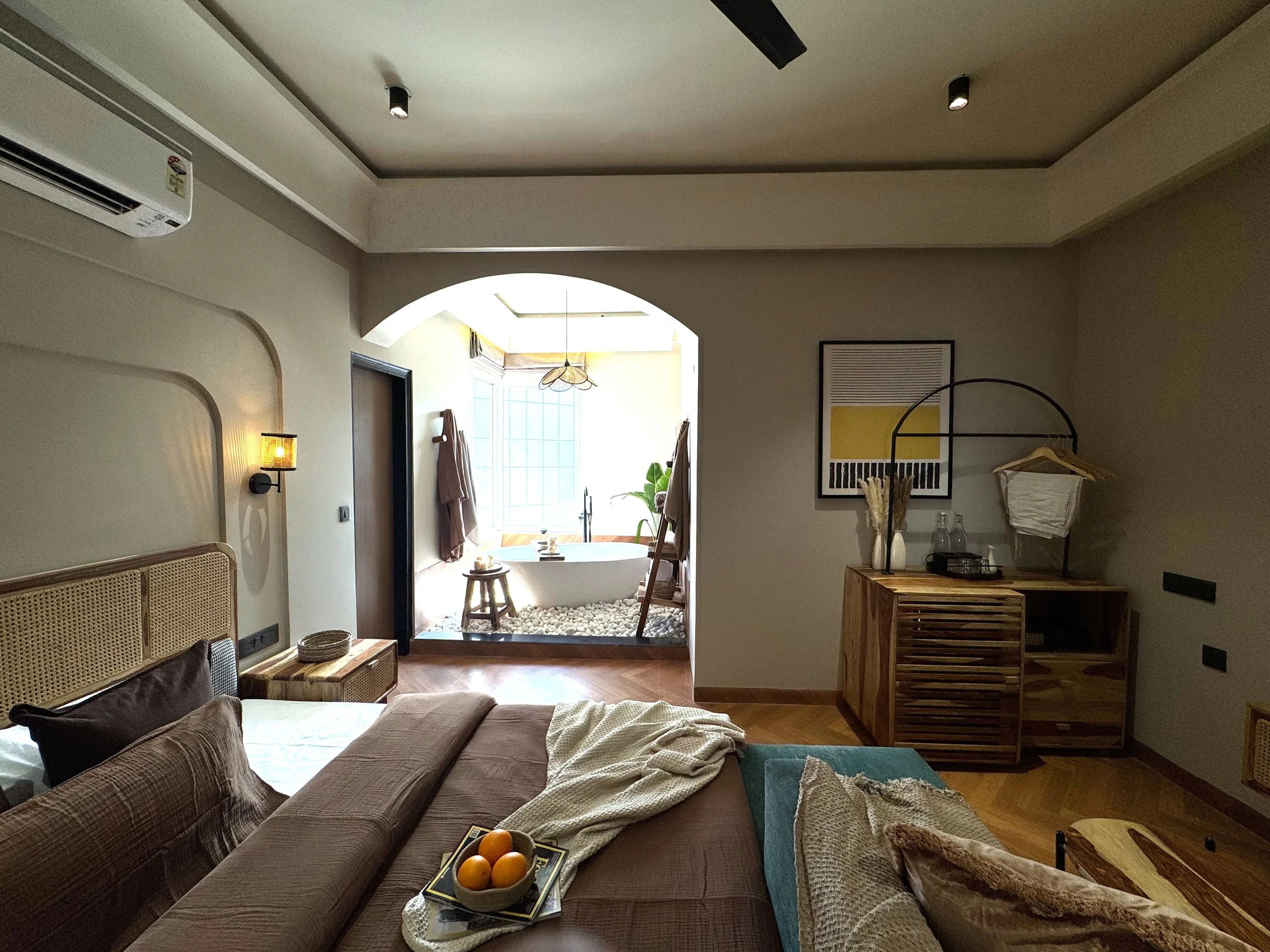 A cozy bedroom with a view into a bright bathroom area. The bedroom features a bed with pillows, a bedside table, and a blanket. The bathroom area has a bathtub, two towels hanging on hooks, and a decorative window with natural light. There is artwork and a wooden cabinet with glass bottles and a hanging rack.