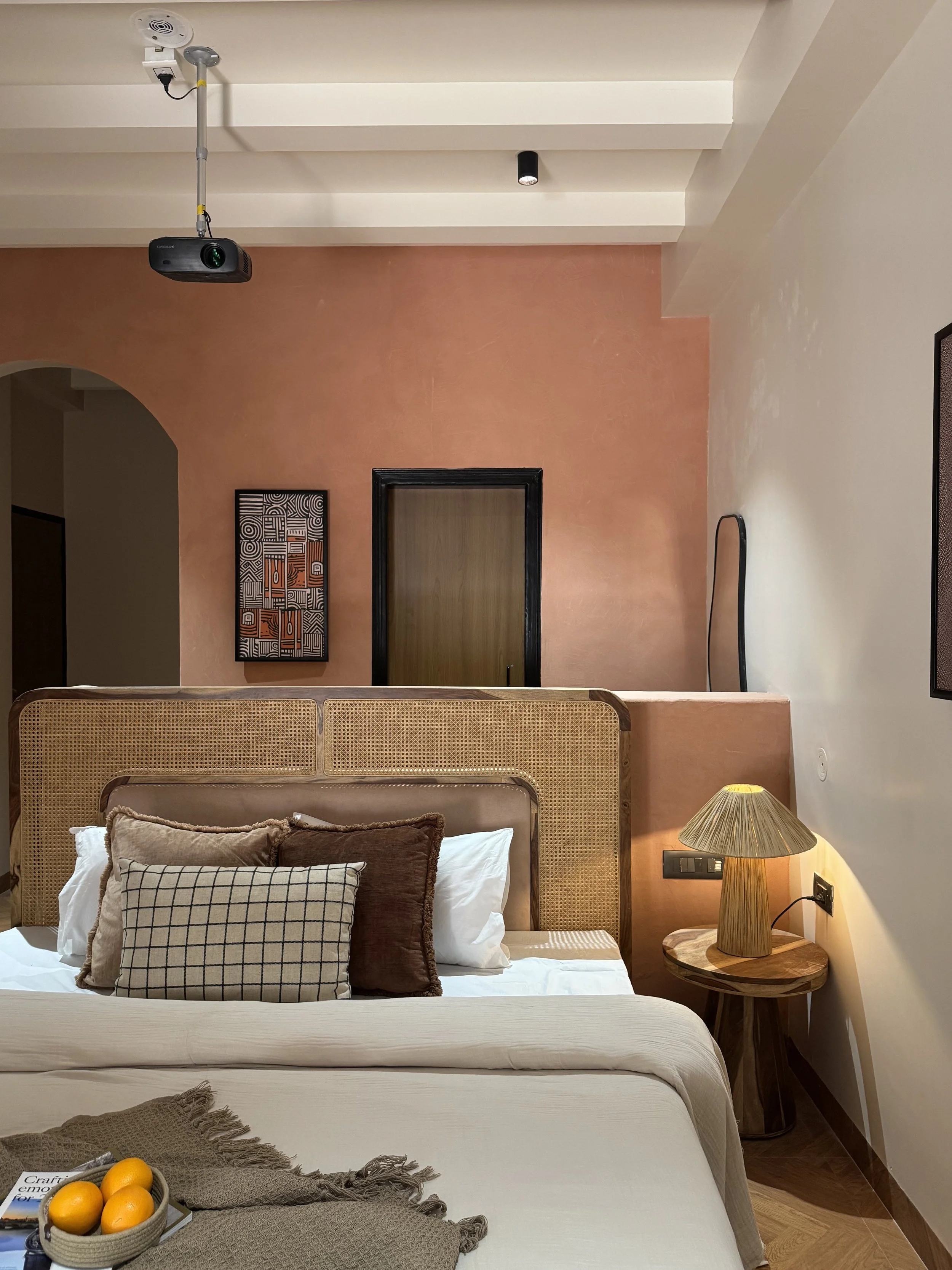 A cozy hotel bedroom with a bed, a side table with a lamp, decorative pillows, a peach-colored wall, and a mounted projector on the ceiling.