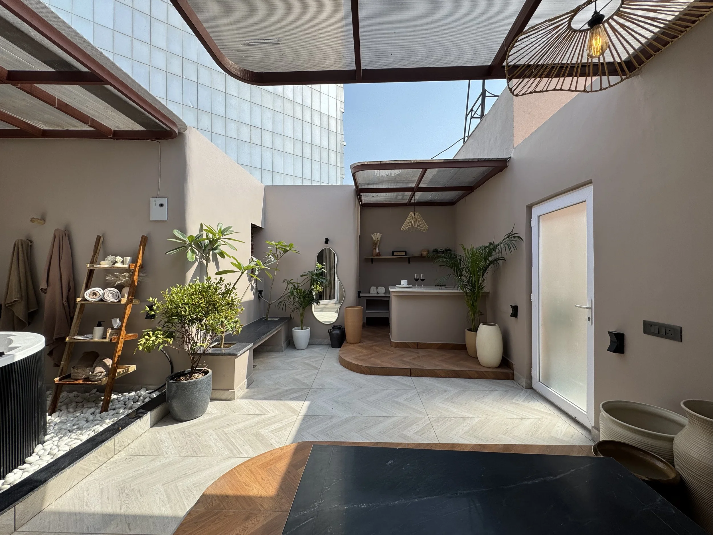 Modern indoor patio with sunlight, potted plants, a bar counter with wine glasses, and decorative elements like a mirror and hanging lamps.