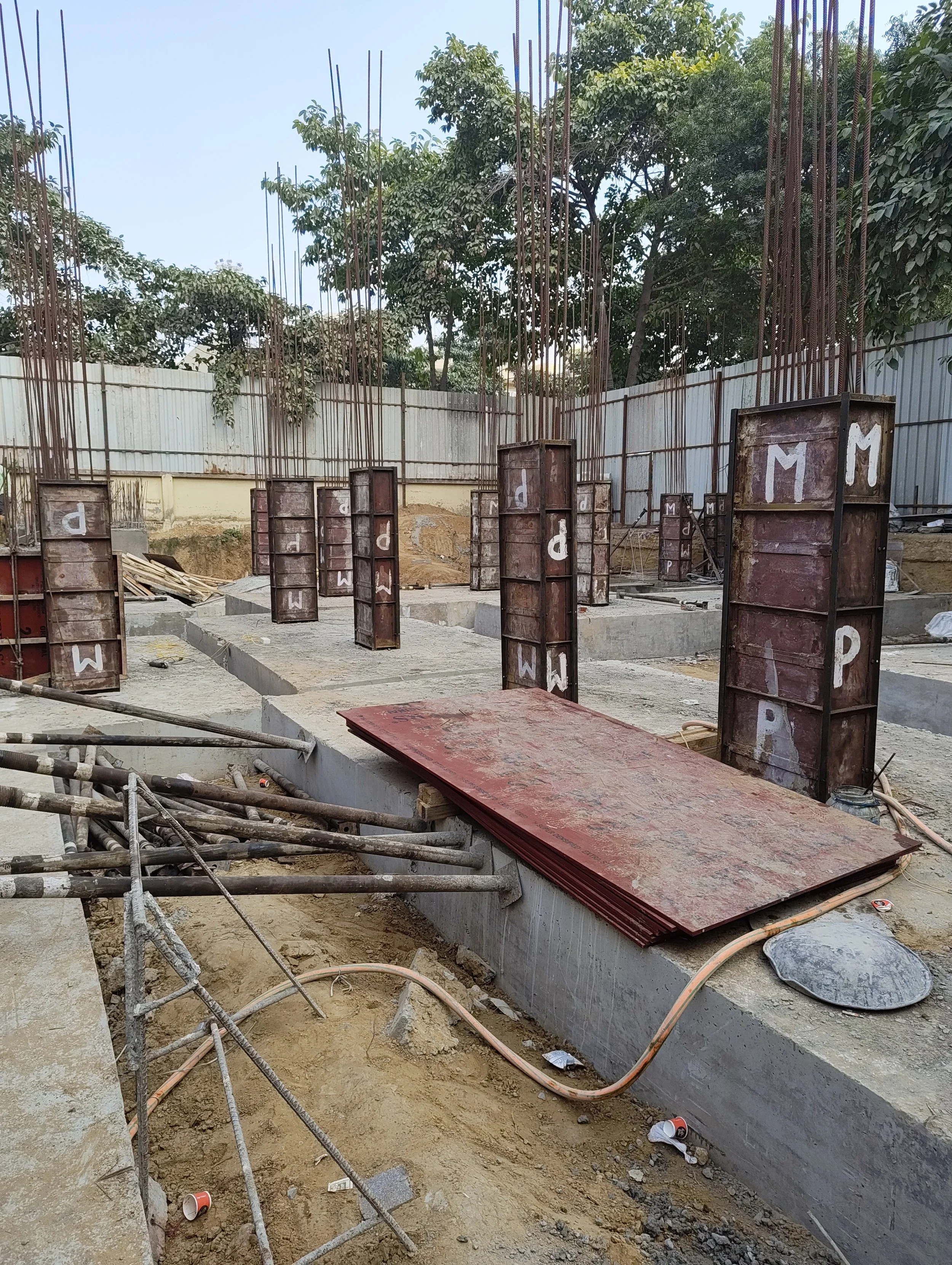 Construction site with steel formwork and rebar for concrete columns. The area is surrounded by a metal fence, with trees visible in the background. Construction materials and tools are scattered around.