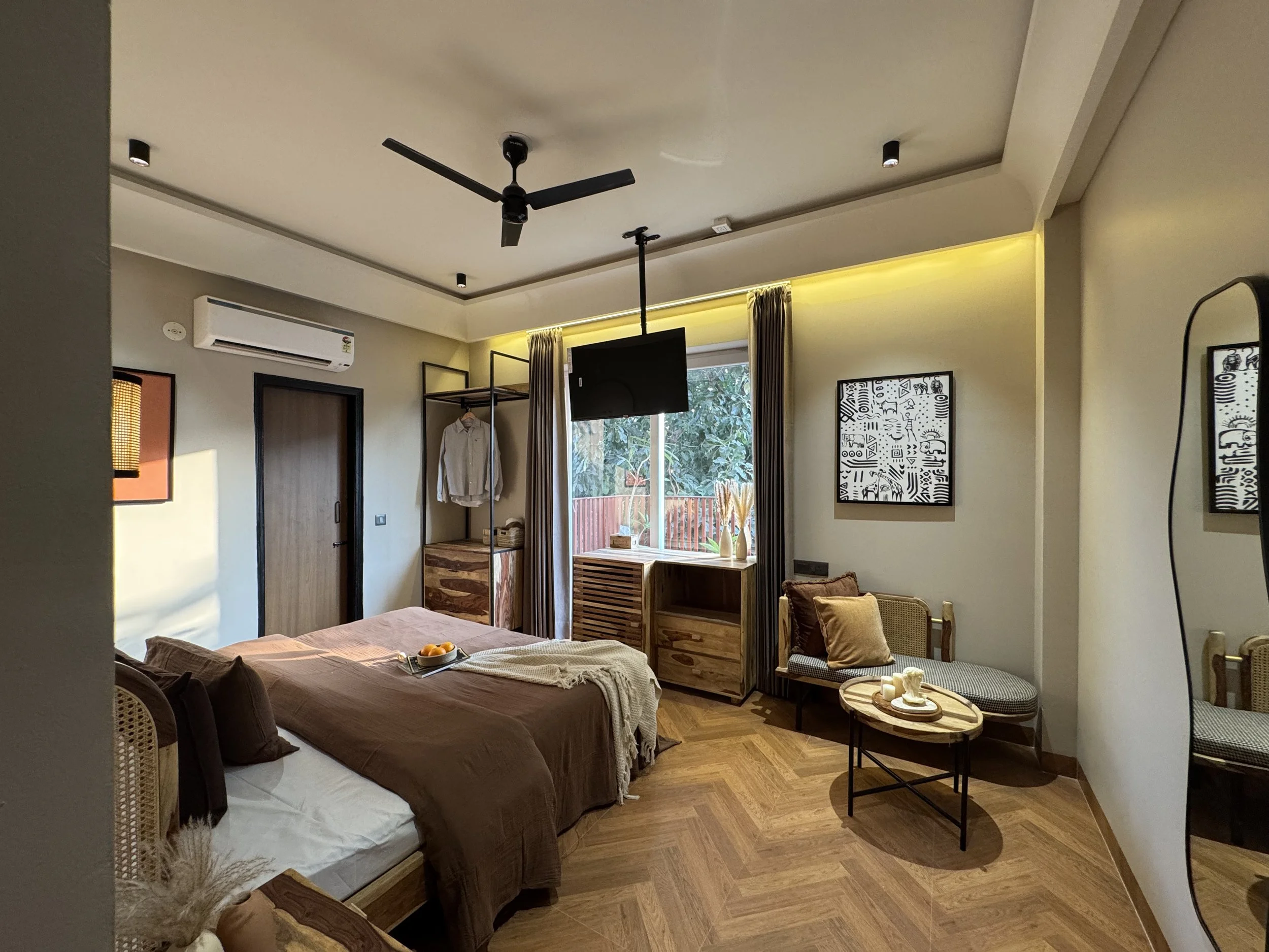 A modern bedroom with a large bed covered in brown and beige bedding, a small sitting area with two chairs and a round table, and a window with curtains showing greenery outside.