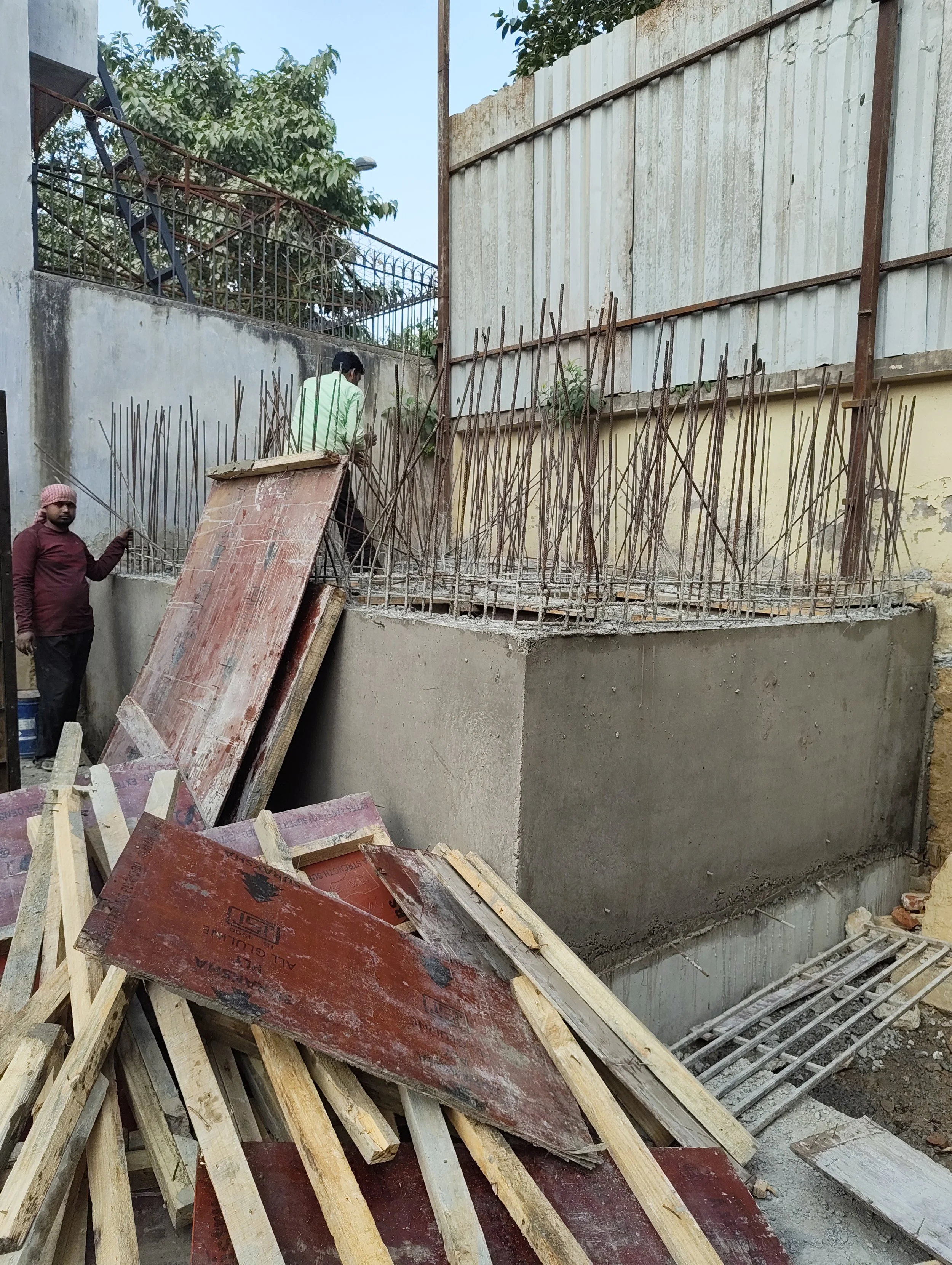 Construction site with two workers installing steel reinforcement bars for a concrete structure, with wooden planks and construction materials in the foreground and a tall fence in the background.