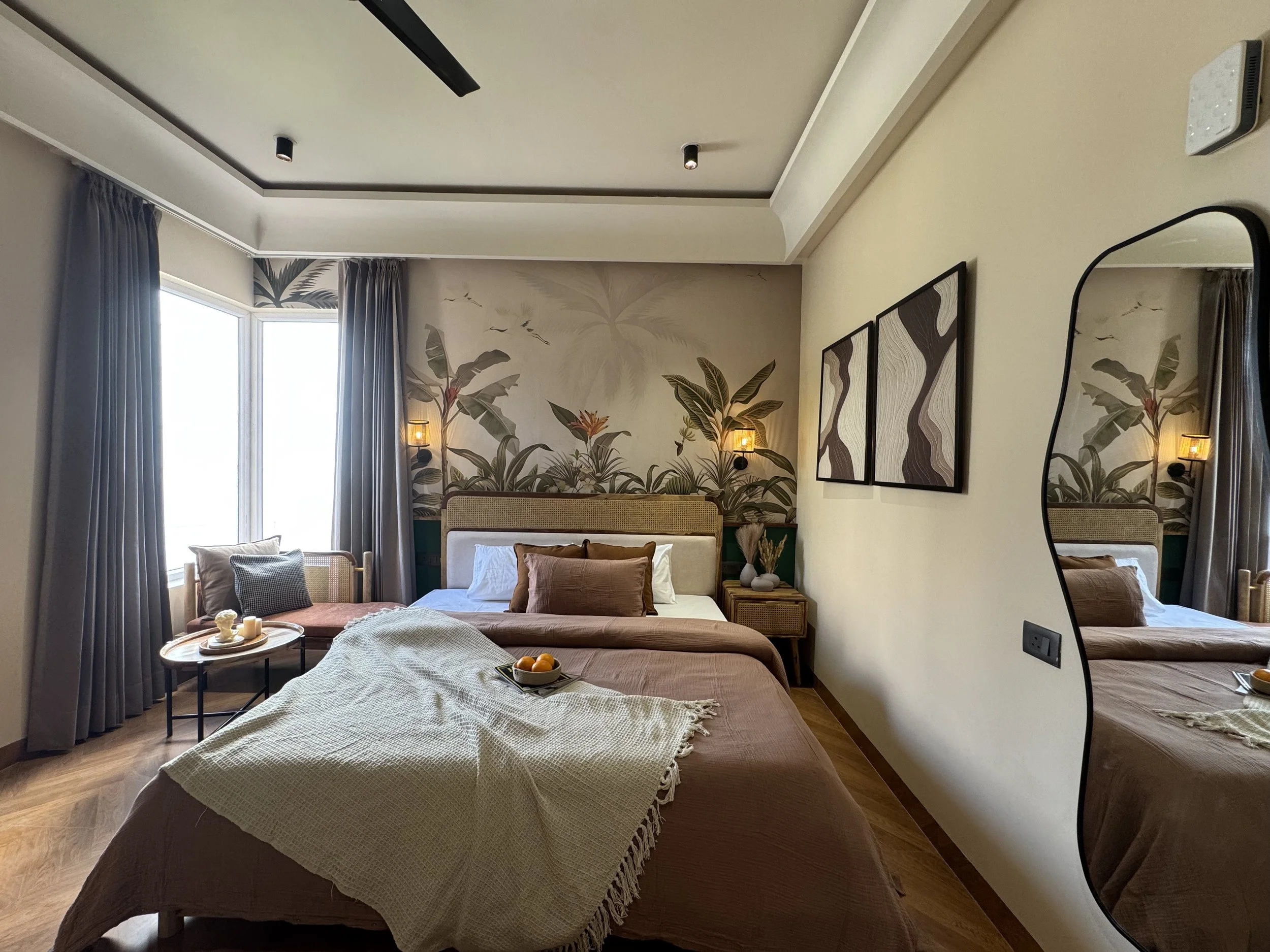 A cozy bedroom with a large bed, decorated with earthy tones and pillows. There's a small round table with snacks and drinks, a sitting area by the window, lush tropical wall art behind the bed, and a large irregularly shaped mirror reflecting part of the room.
