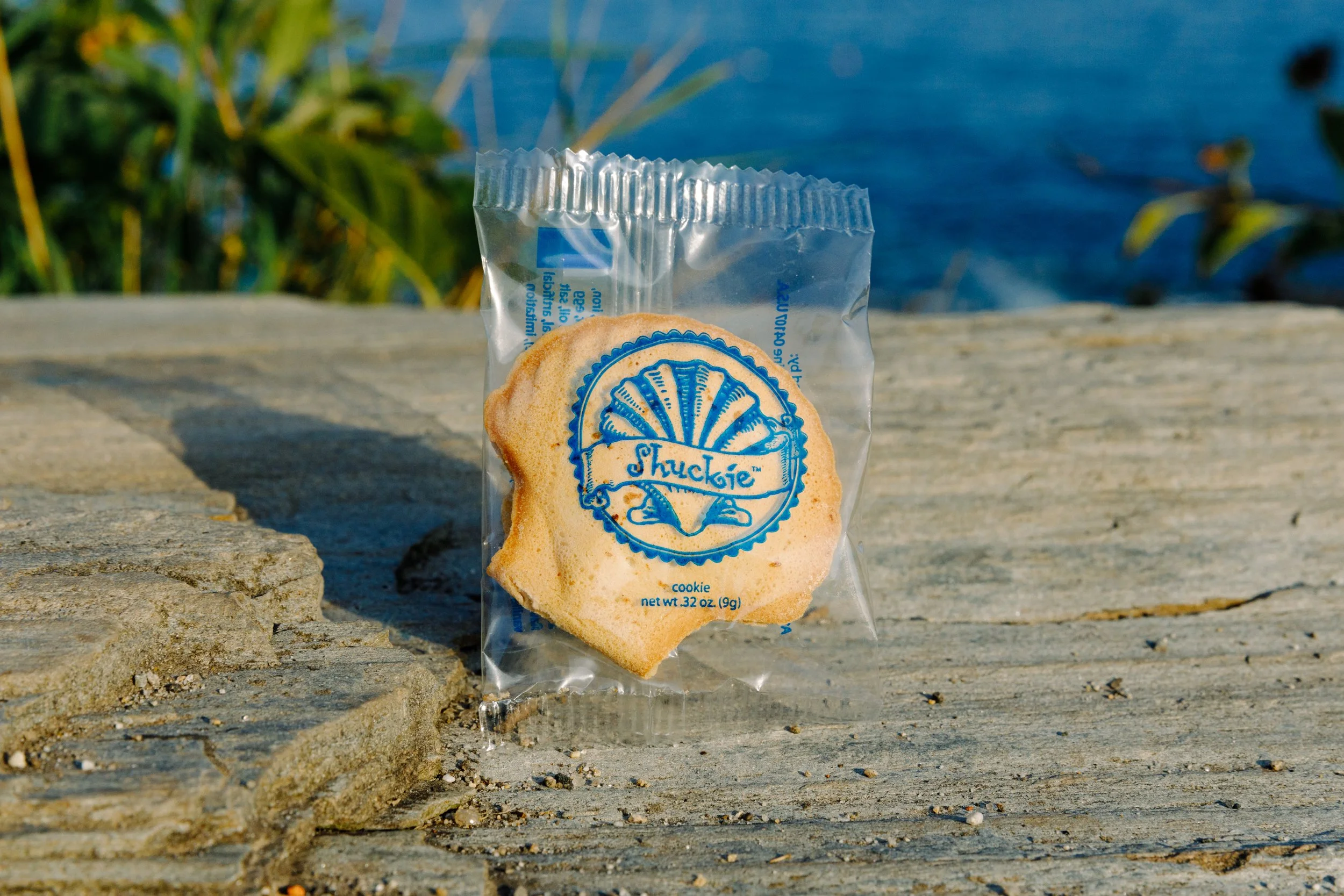 A packaged cookie with the brand name 'Shuckie' printed on the wrapper, placed on a wooden surface outdoors near water and green vegetation.