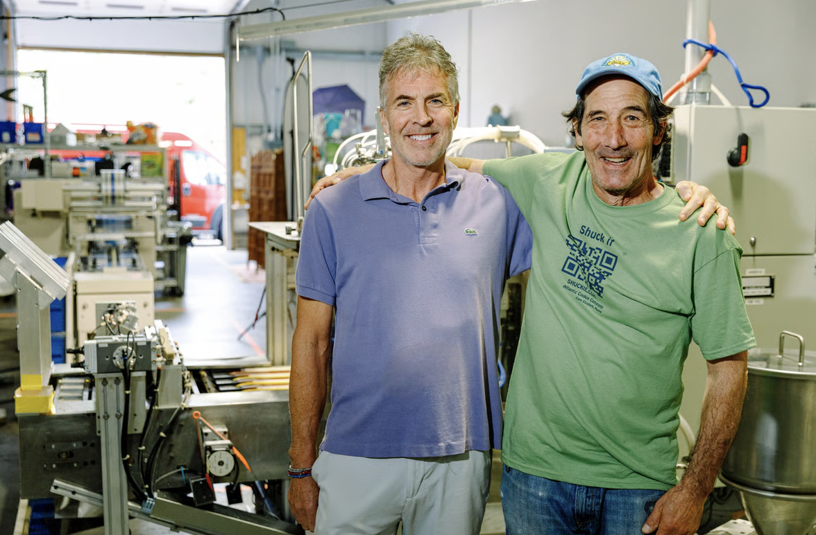 Two men smiling and standing with arms around each other in a workshop, surrounded by industrial machinery and equipment.