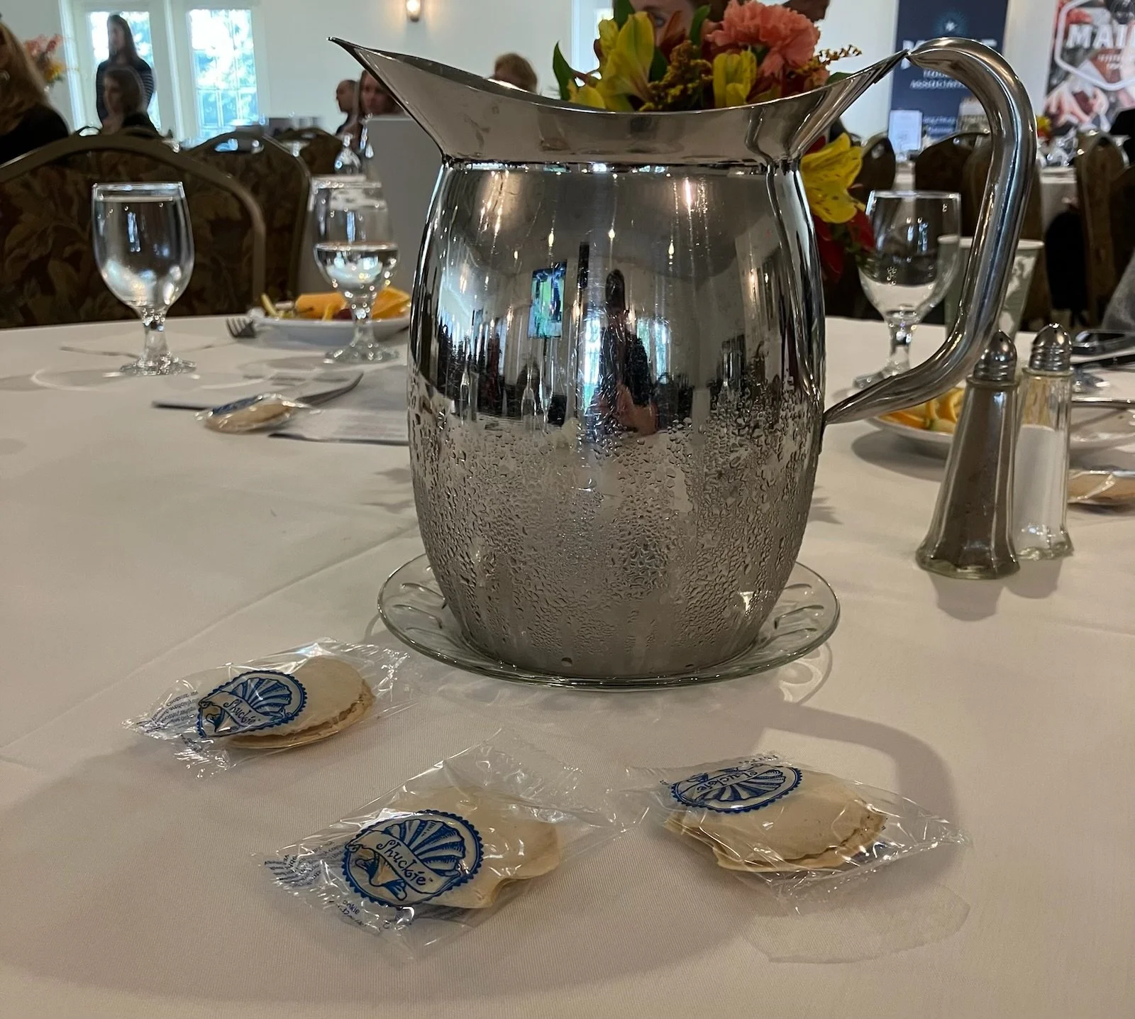 A table set with a large silver pitcher of water, three wrapped mints in plastic wrappers with a blue logo, several empty wine glasses, and a tray of sliced fruit in the background. A floral centerpiece is also visible.