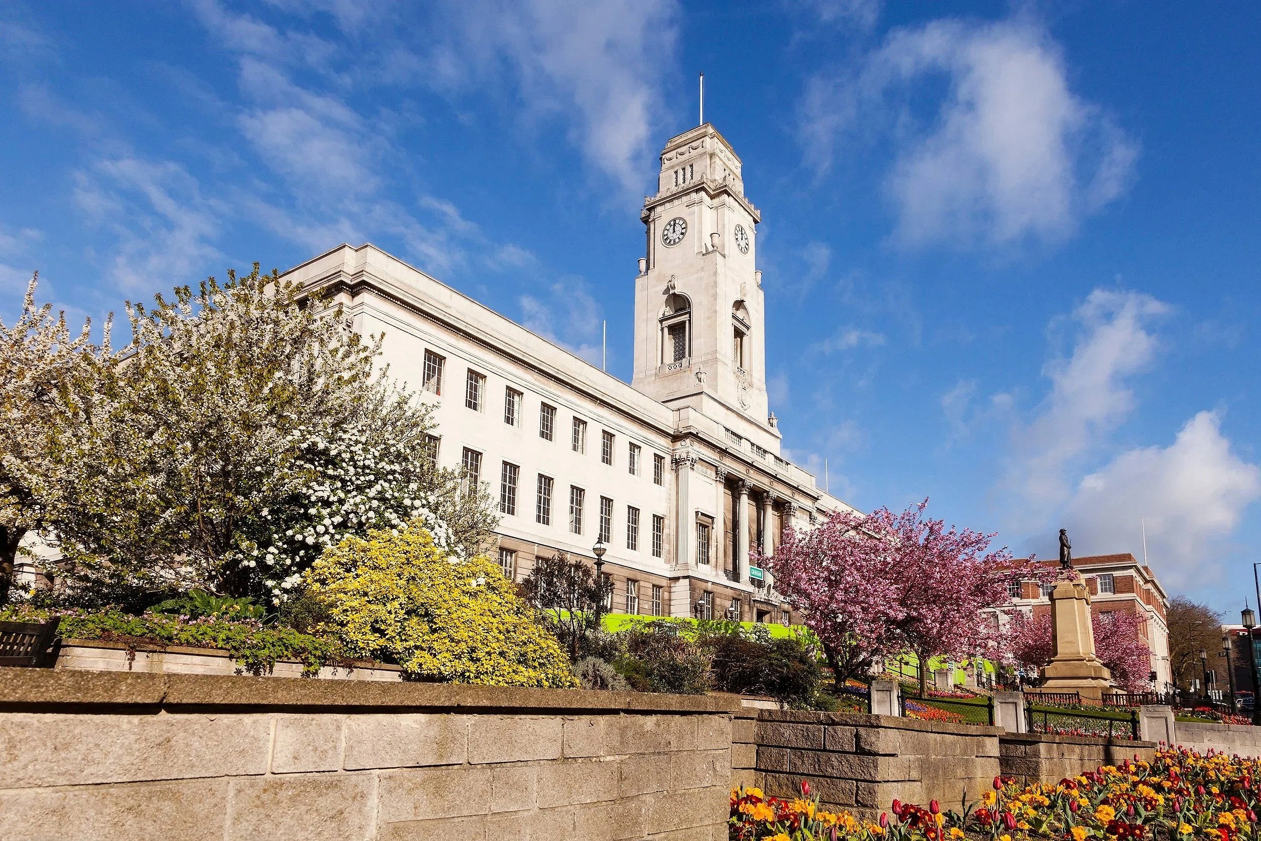 A historic white building with a clock tower under a blue sky with scattered clouds, surrounded by blooming cherry blossom trees and colorful flowers.