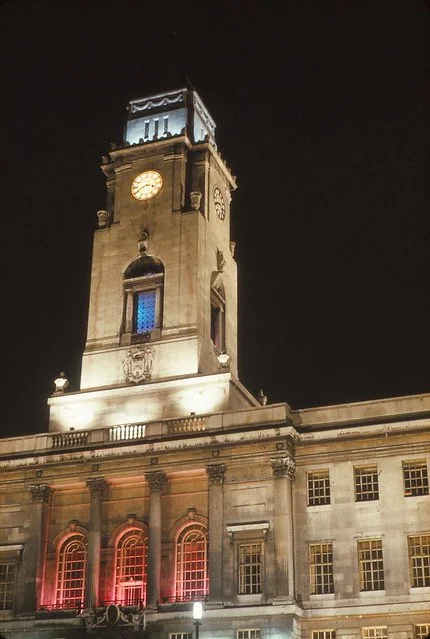 A tall historical clock tower illuminated at night, attached to a grand stone building with arched windows and decorative columns. Barnsley Town Hall at night