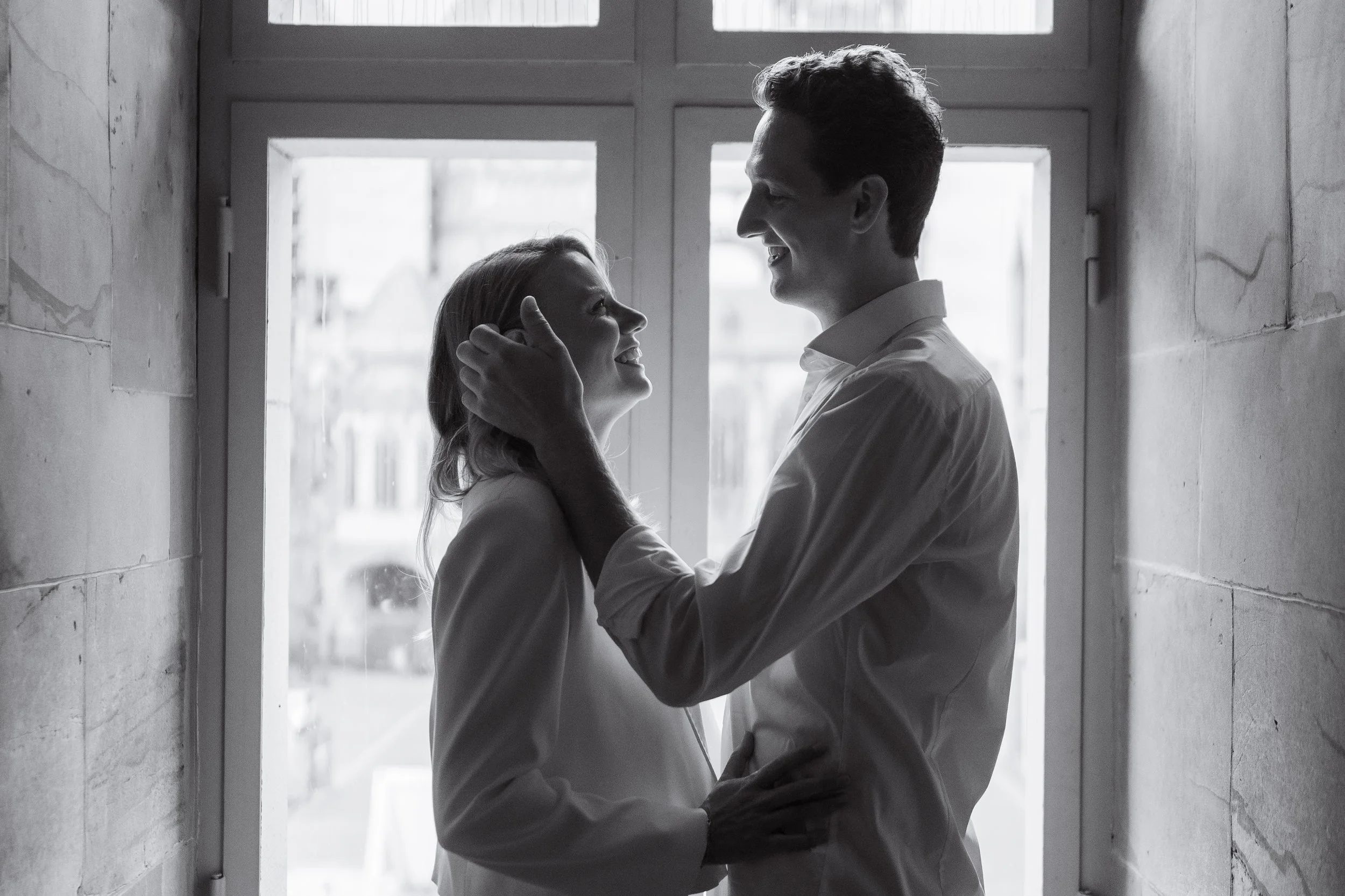 Ein glückliches Paar in Schwarz-Weiß, das in einem engen Flur vor einem Fenster steht, sie lächeln sich an.