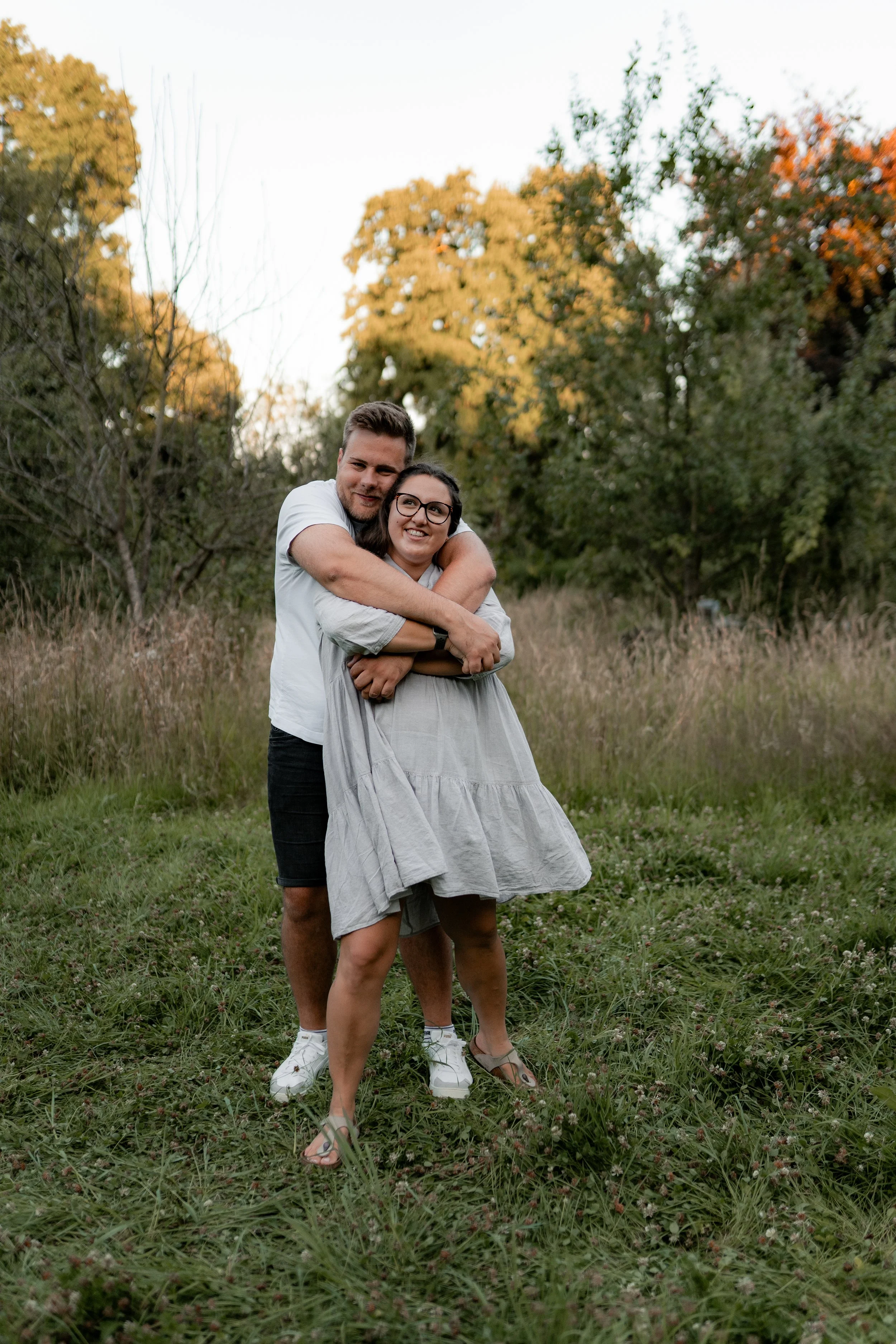 Ein Paar umarmt sich lachend in einem grünen Feld im Freien mit Bäumen im Hintergrund bei Sonnenuntergang.