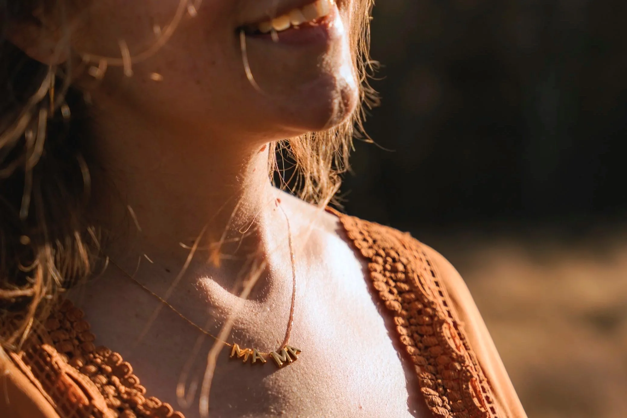 Nahaufnahme eines Frauenhalses mit goldener Halskette mit den Buchstaben 'MAMA' und 'MOM' im Sonnenlicht, trägt einen braunen, bestickten Kleidungsstoff.