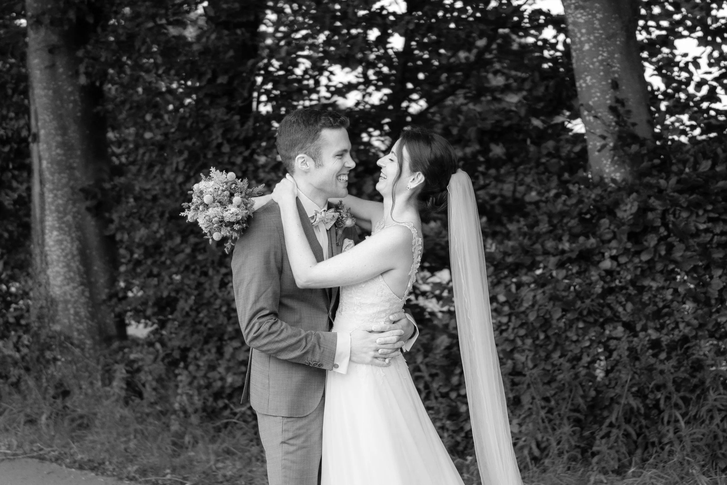 Braut und Bräutigam bei ihrer Hochzeit im Freien, lachend und umarmend, mit einem Baum und Laub im Hintergrund, in Schwarzweiß.