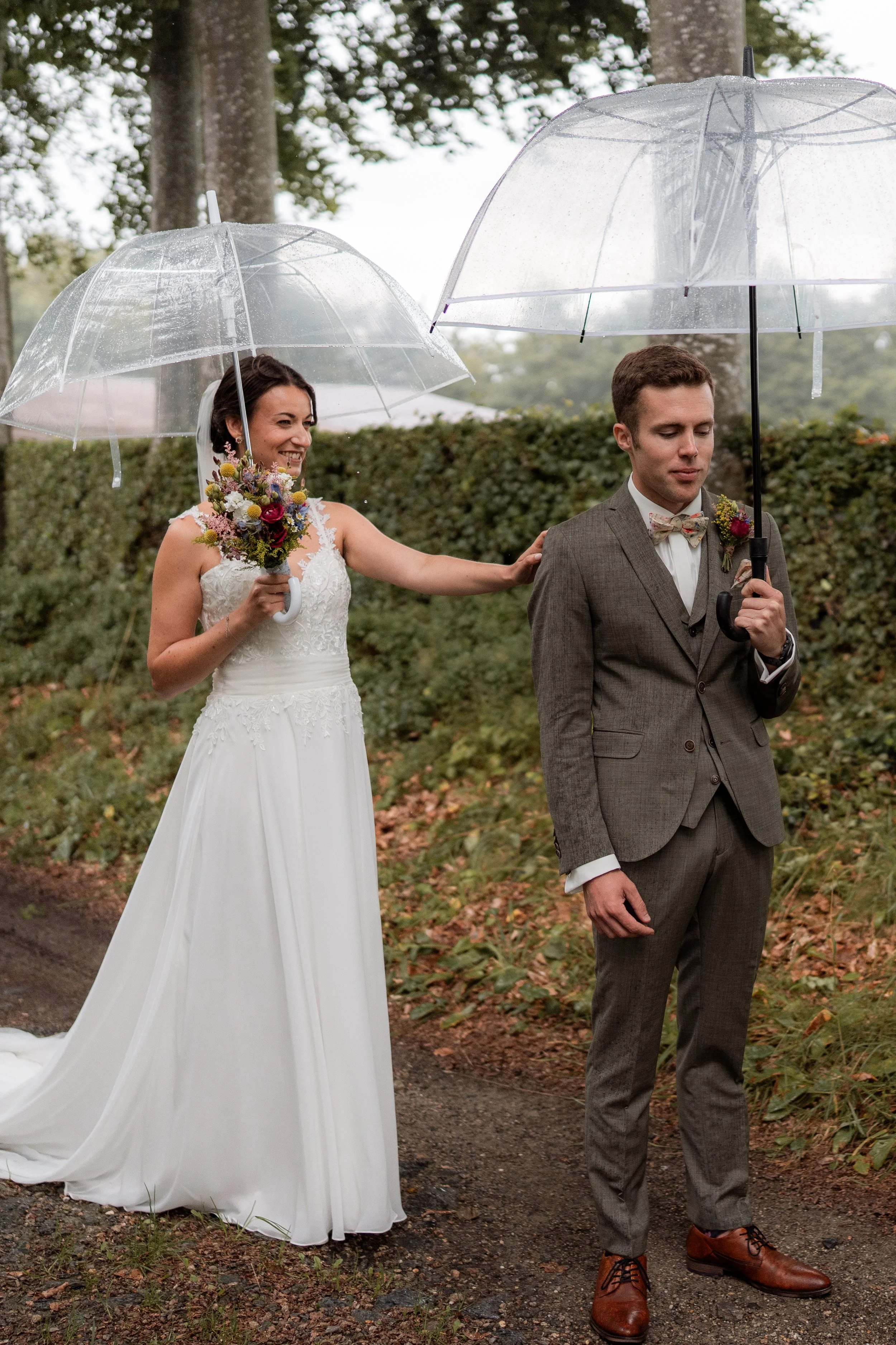 Ein Brautpaar steht bei Regen im Freien, die Braut in einem weißen Kleid mit Blumenkette und der Bräutigam in einem grauen Anzug, beide halten transparente Regenschirme.
