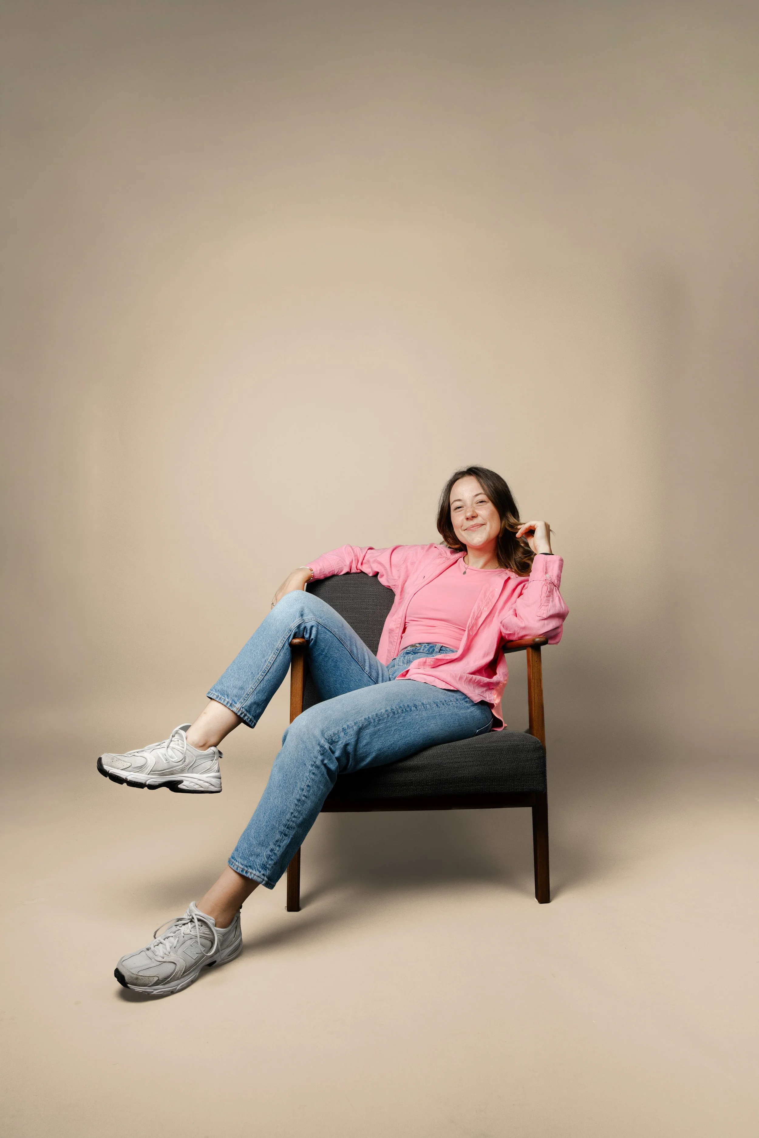 Junge Frau sitzt lässig auf einem Stuhl, trägt pinkes Oberteil, Jeans und Sneakers, lächelt.