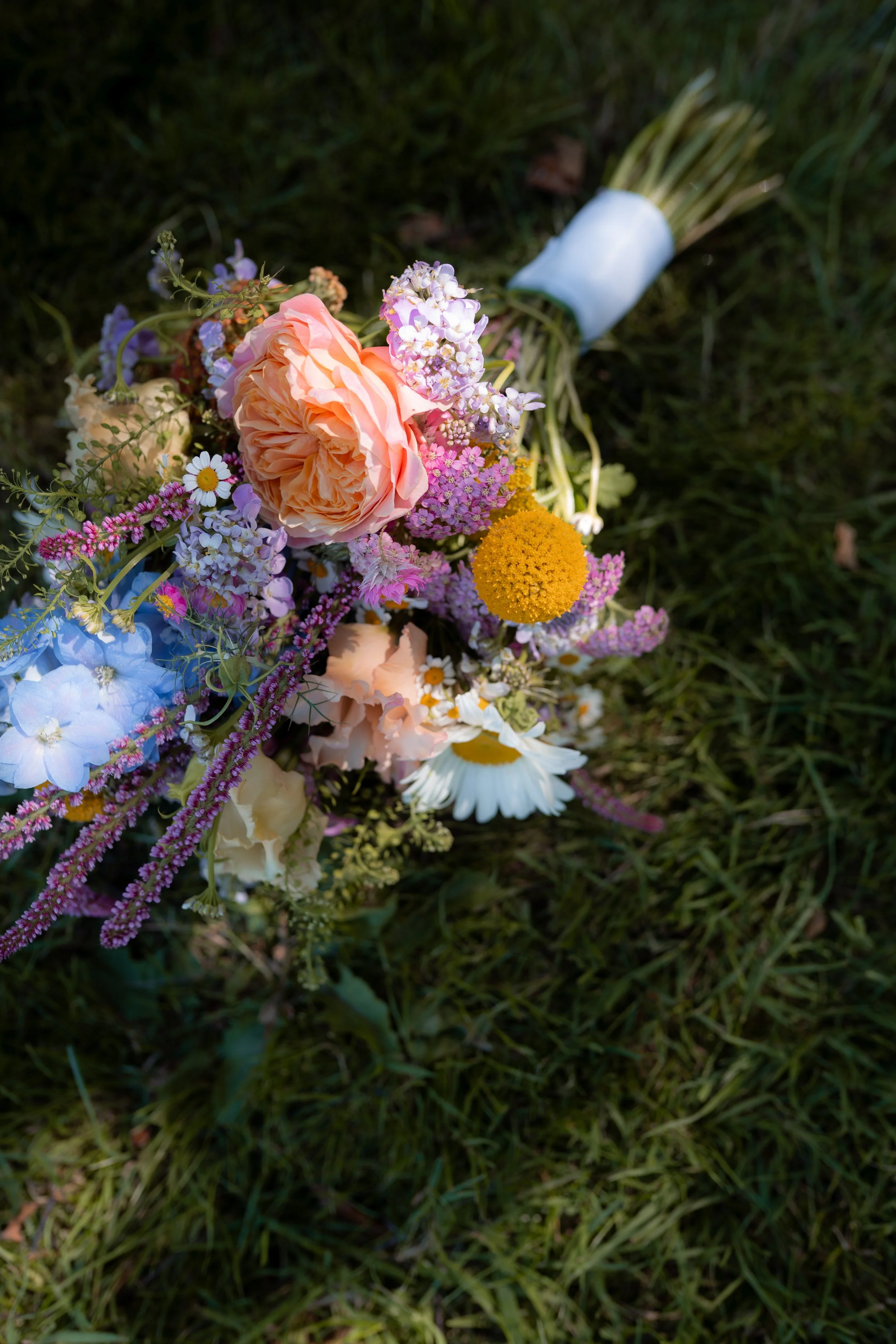 Ein bunter Blumenstrauß mit Rosen, Gänseblümchen, Fetthenne und anderen Blumen liegt auf Gras.