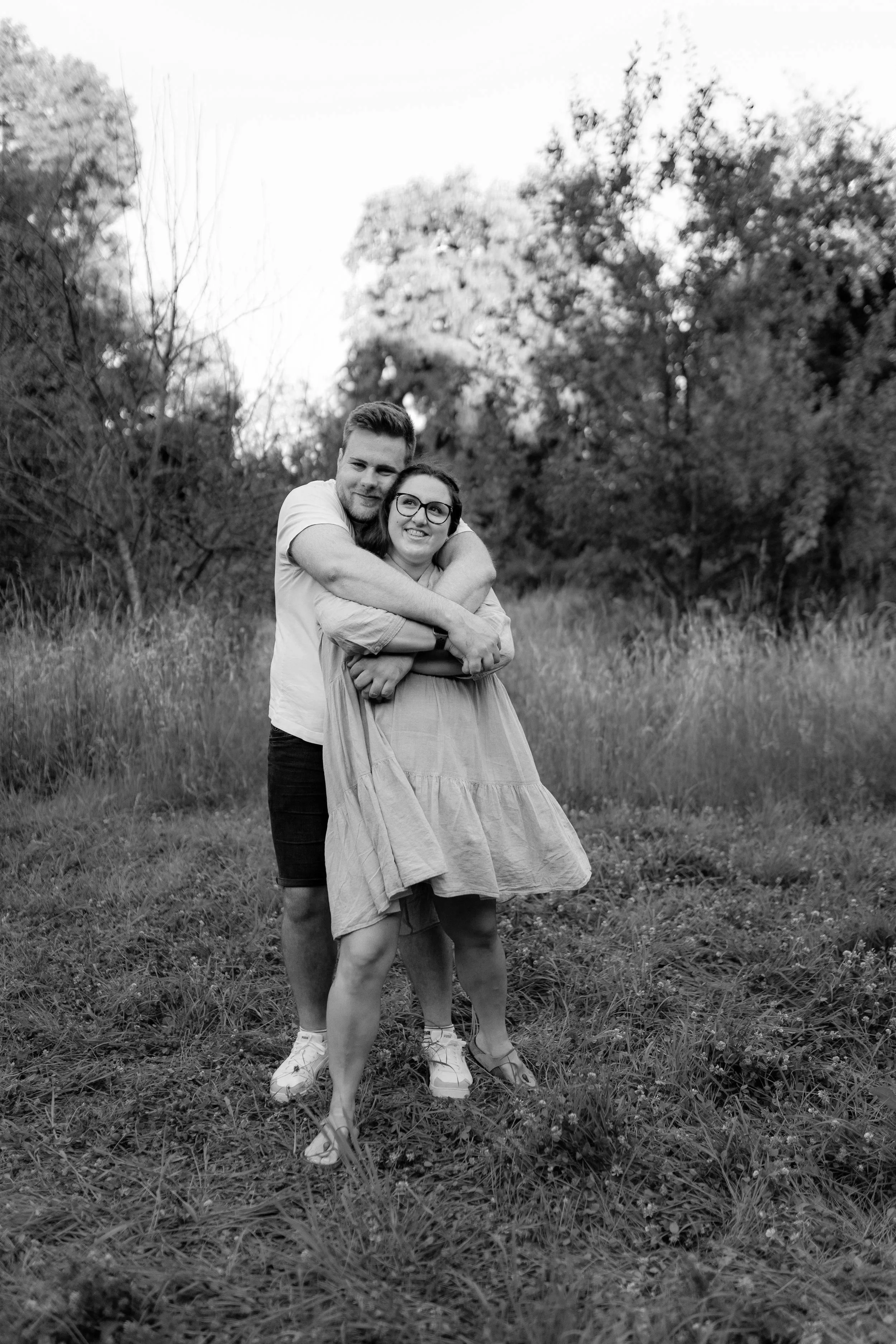 Ein lachendes Paar, umarmt im Freien, auf einer Wiese mit Bäumen im Hintergrund, in schwarz-weiß