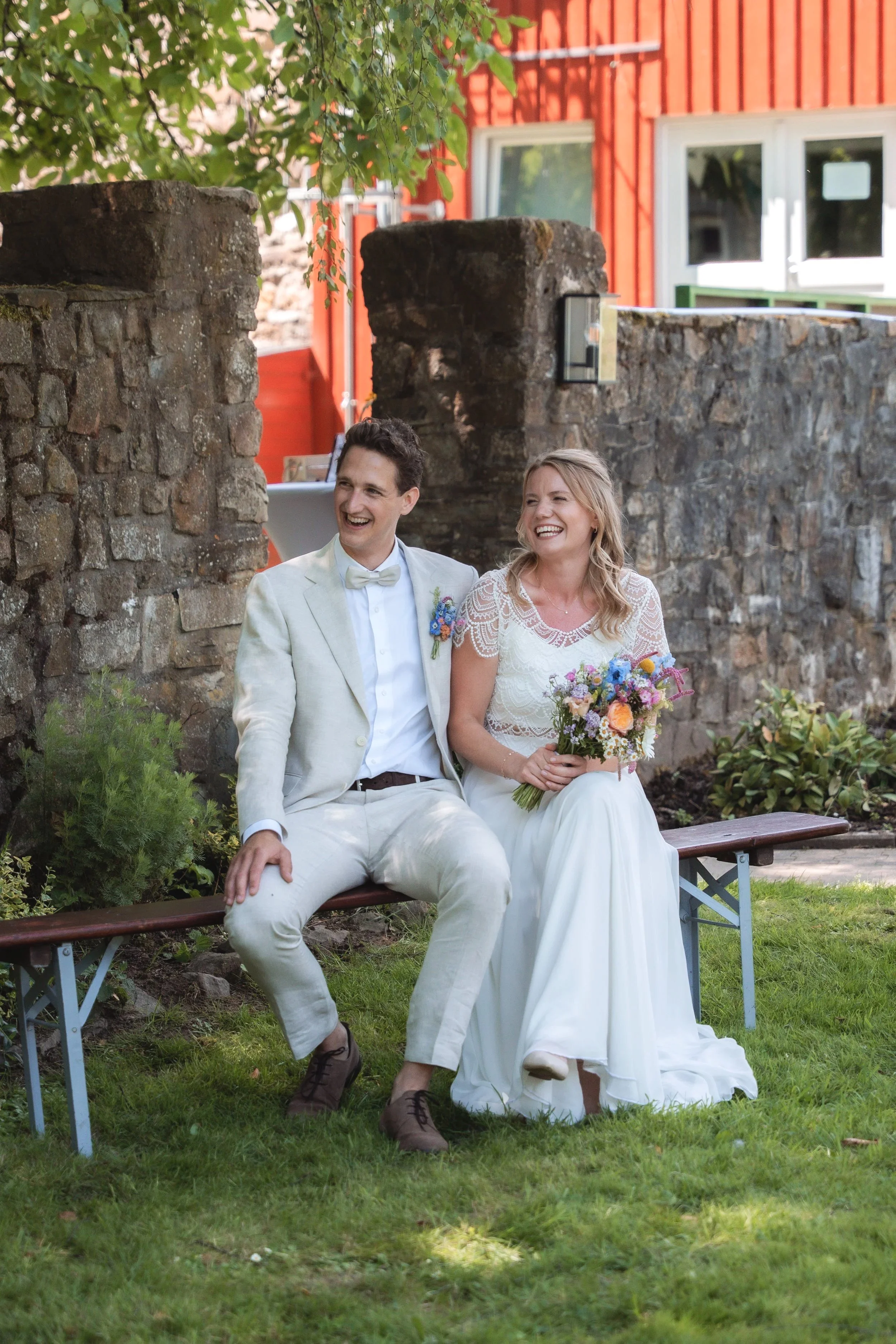 Ein kürzlich vermähltes Paar sitzt lachend auf einer Bank im Garten, umgeben von Grün und einem alten Steinmauer, mit einem roten Haus im Hintergrund.