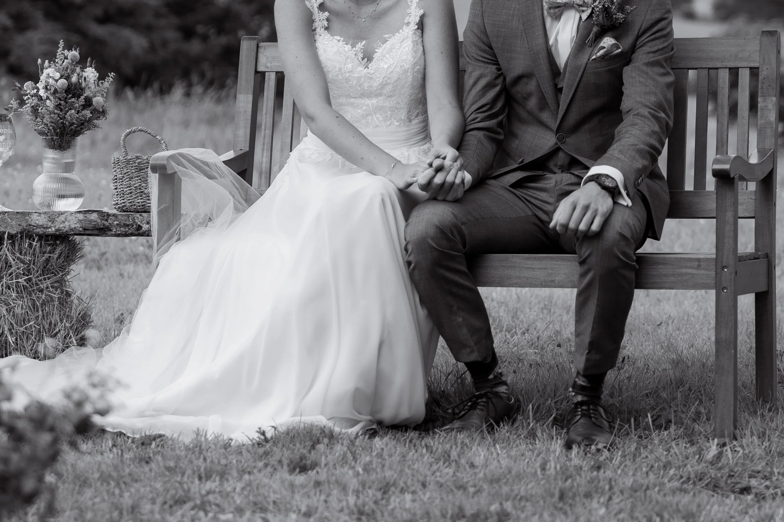 Ein Brautpaar sitzt auf einer Holzbank im Freien, die Braut trägt ein weiße Brautkleid, der Bräutigam einen grauen Anzug, sie halten sich an den Händen, neben ihnen ein Tisch mit Blumen und einem Korb.