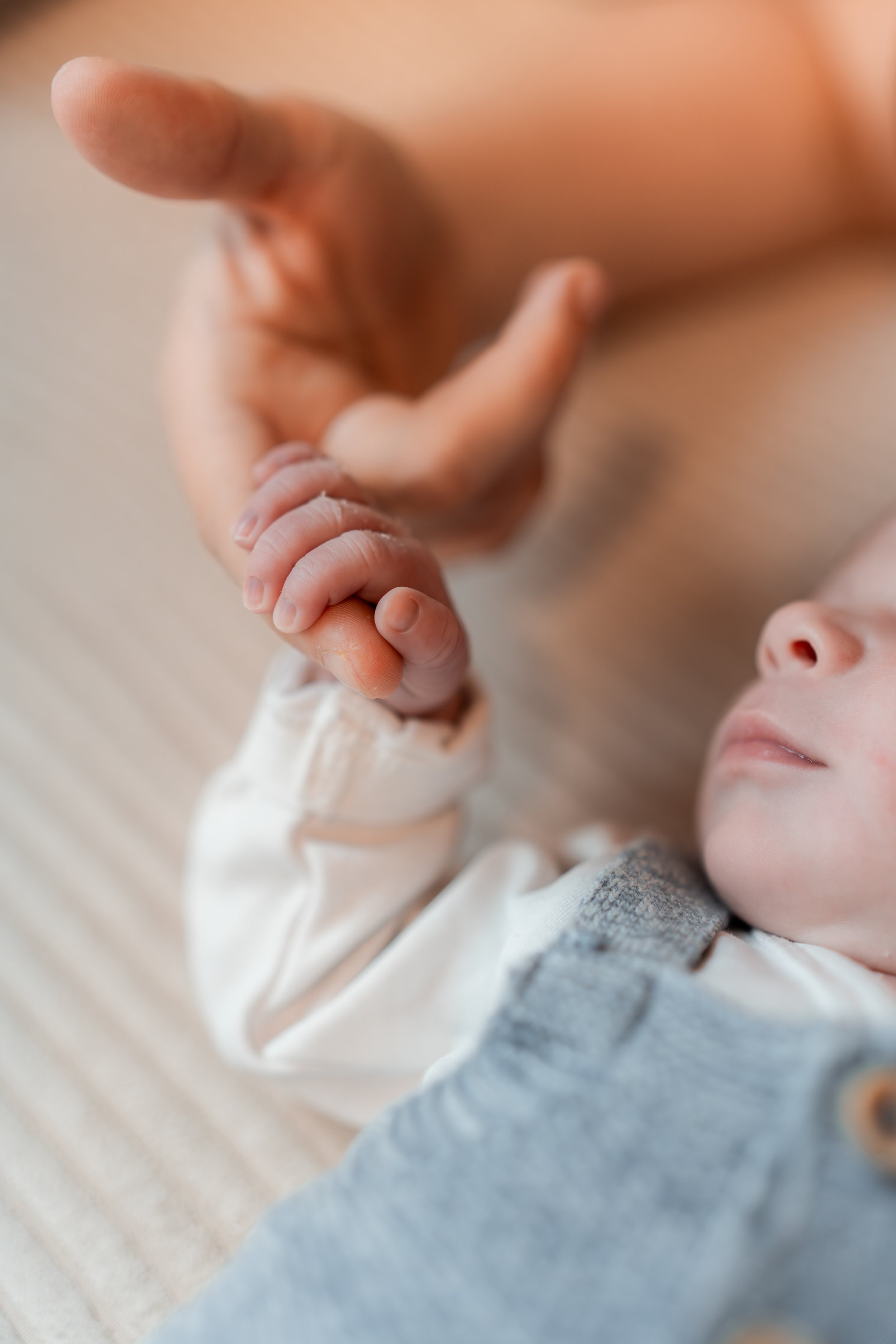 Ein Baby liegt auf dem Boden und hält die Hand eines Erwachsenen. Das Baby ist entspannt und schläft leicht, während der Erwachsene sanft die Hand hält.