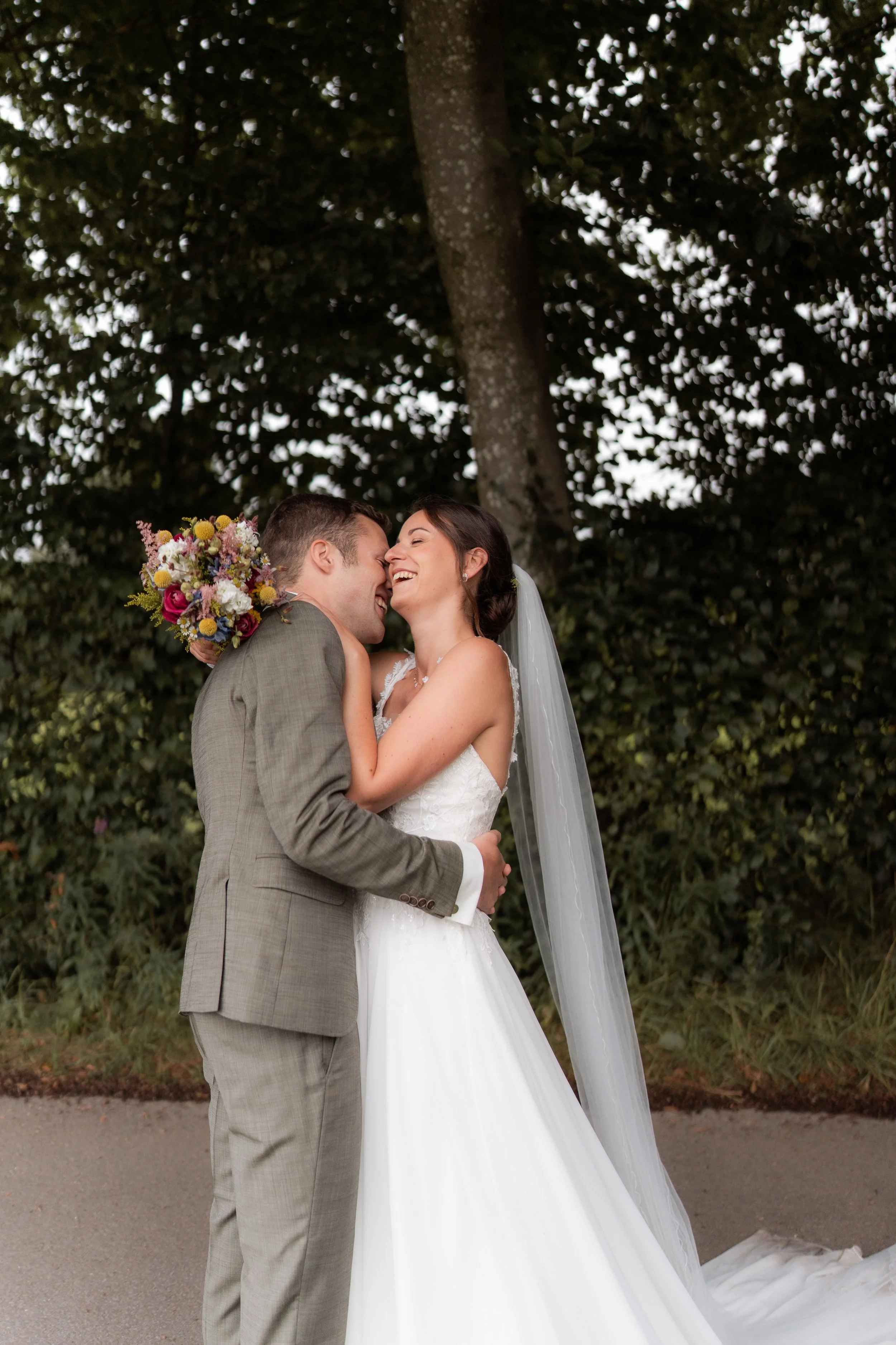 Ein Brautpaar bei der Hochzeit, umarmt sich lächelnd vor einem Baum im Freien.