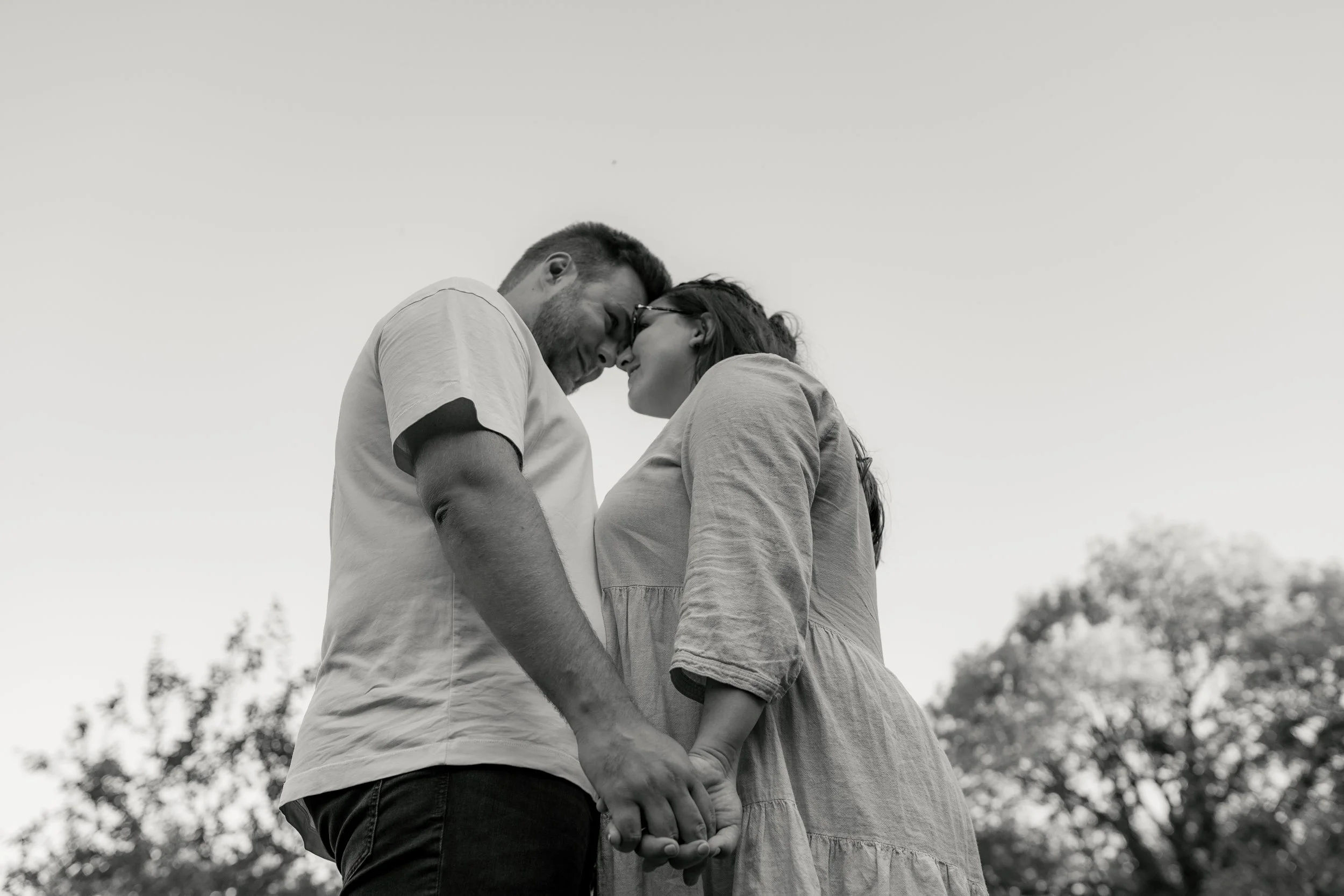 Ein Paar steht im Freien und küsst sich, während sie Händchen halten, umgeben von Bäumen im Hintergrund.