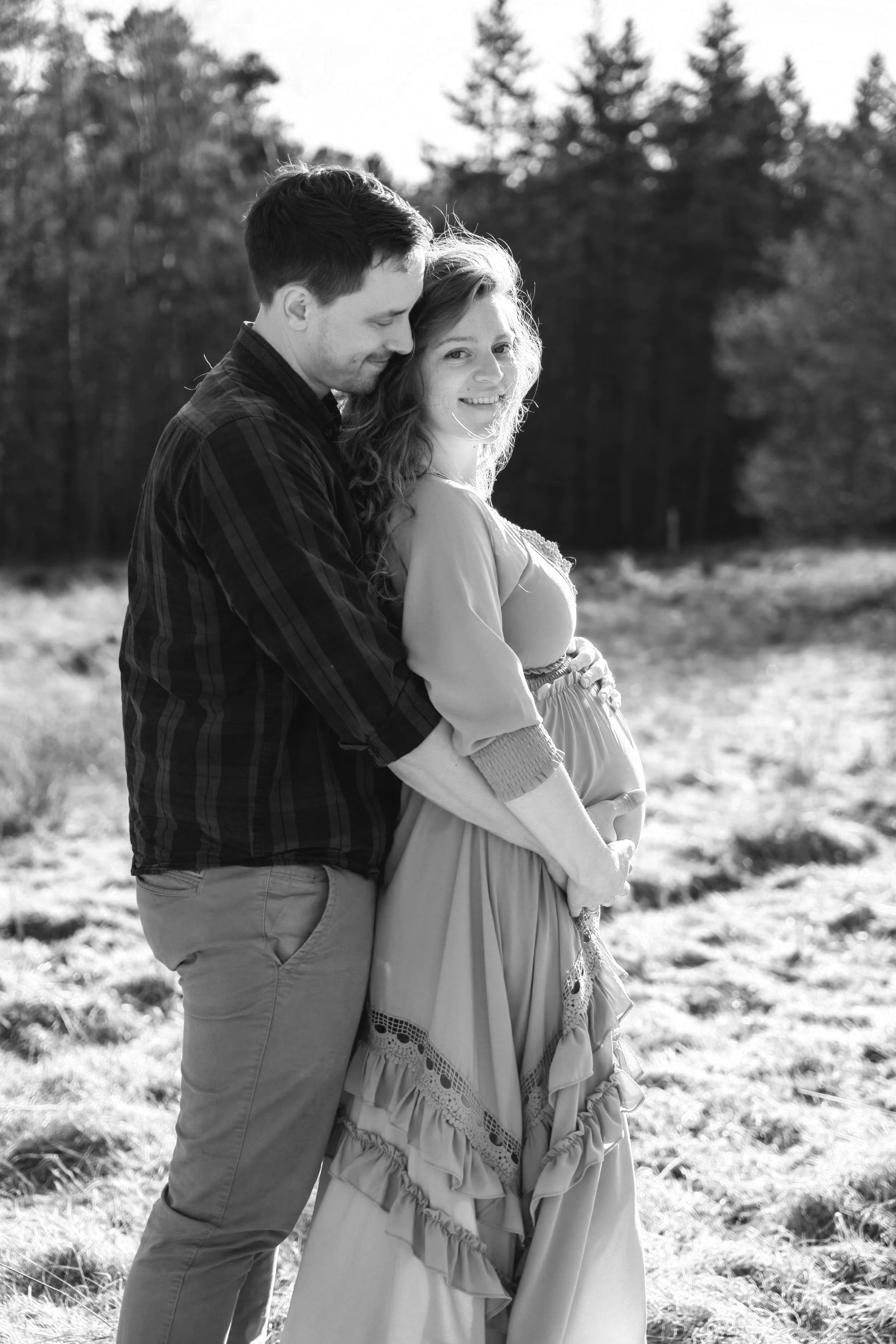 Ein Paar, das draußen in einem Feld steht, romantisch umarmt, die Frau scheint schwanger zu sein, im Schwarz-Weiß-Format.