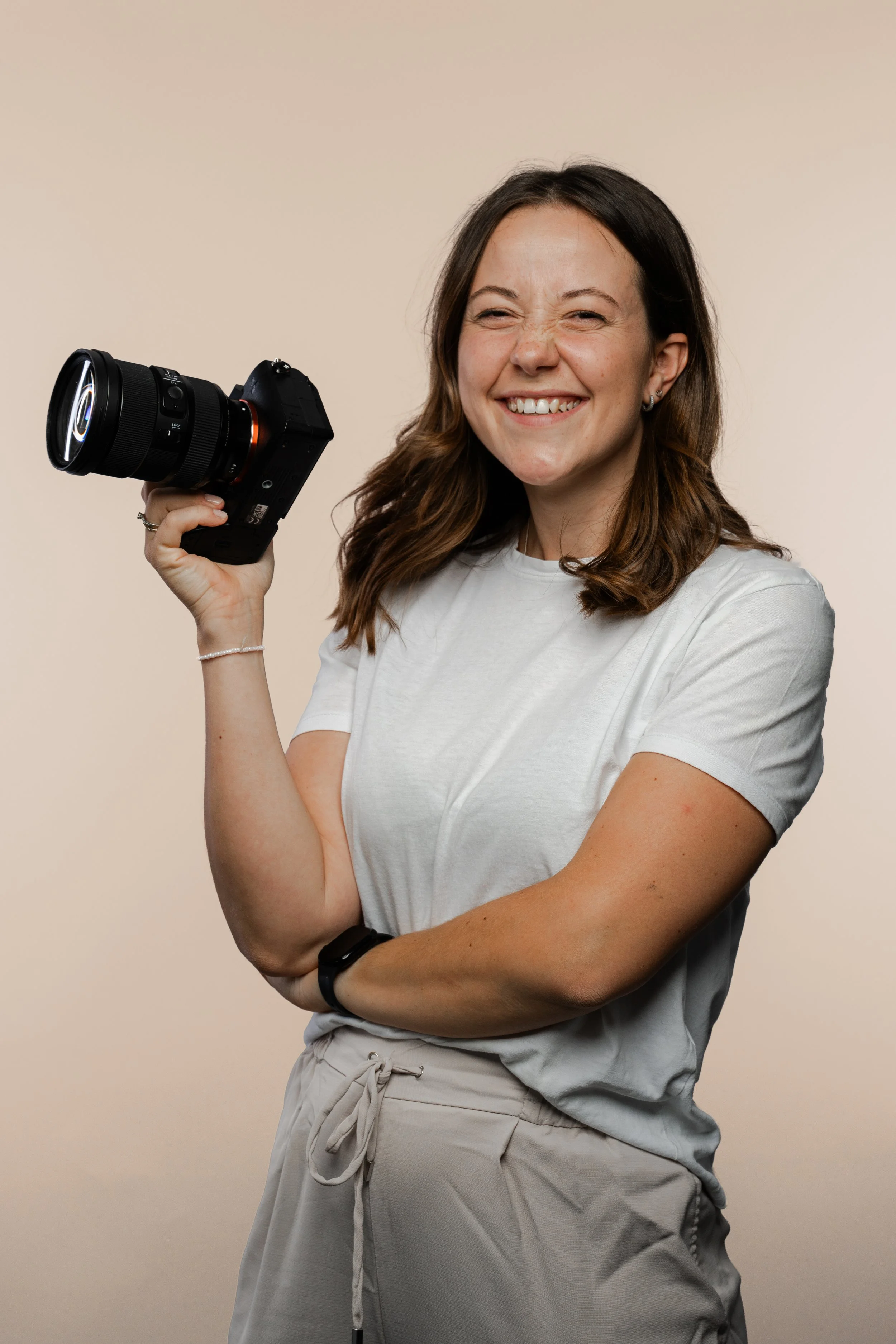 Junge Frau mit braunen Haaren, weißen T-Shirt, Lächeln und einem Kameragehäuse in der Hand vor hellem Hintergrund.