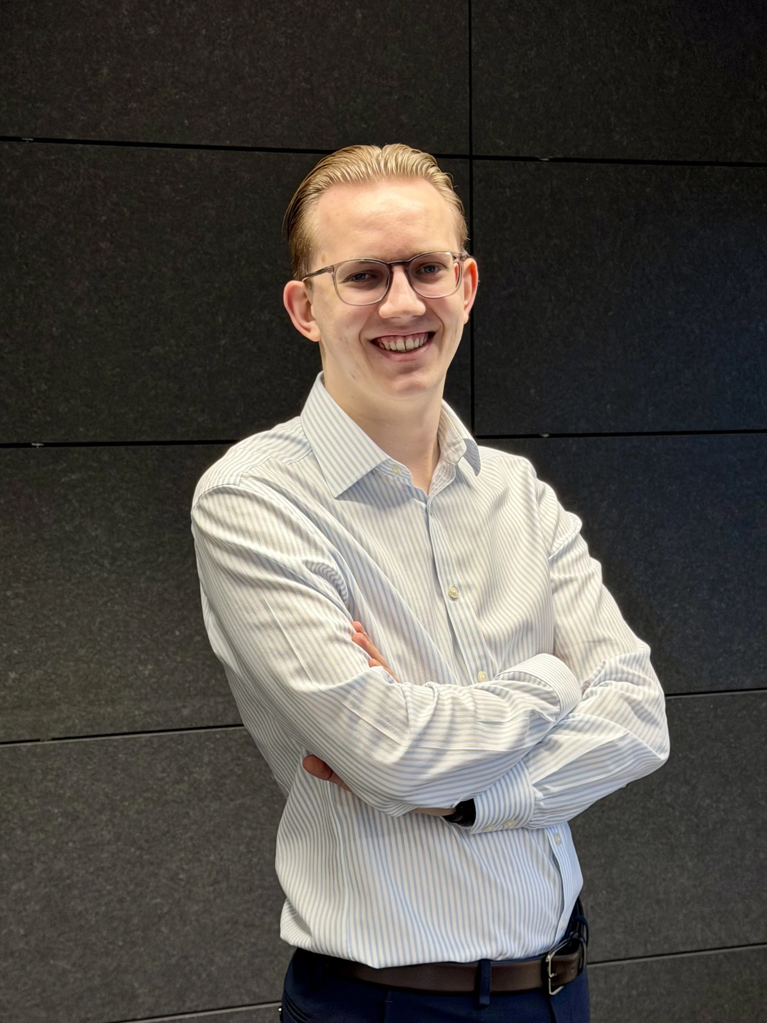 Junge Mann mit Brille, lächelnd, trägt ein weißes Hemd mit Streifen, steht vor einer schwarzen Wand. REM TRANSACTION GmbH