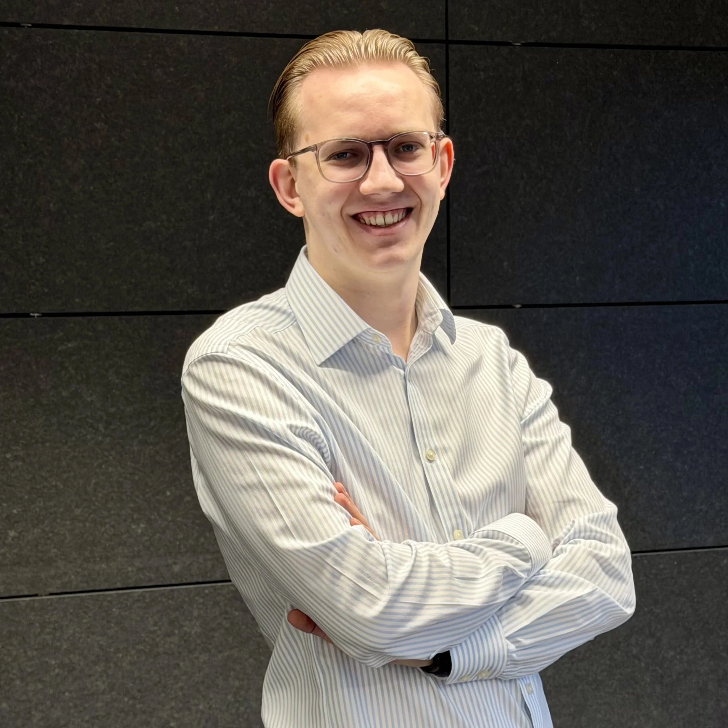 Ein junger Mann mit Brille und einem weißen, gestreiften Hemd steht vor einer schwarzen Wand. Er lächelt und hat die Arme verschränkt. Ben Berger, Praktikant der REM TRANSACTION GmbH.