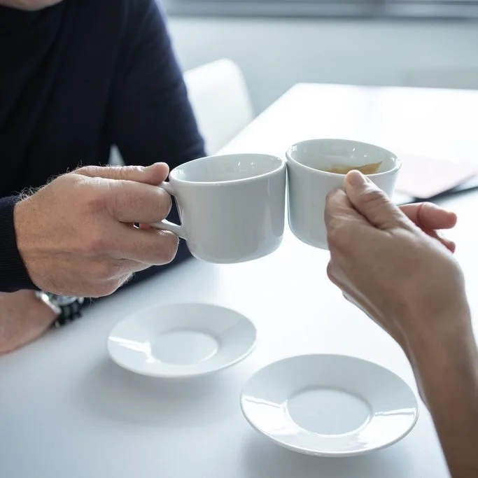 Menschen stoßen mit Kaffeetassen an, auf einem weißen Tisch mit Untertassen, im Hintergrund ein Fenster. Symbolbild für M&A Beratung. REM TRANSACTION GmbH