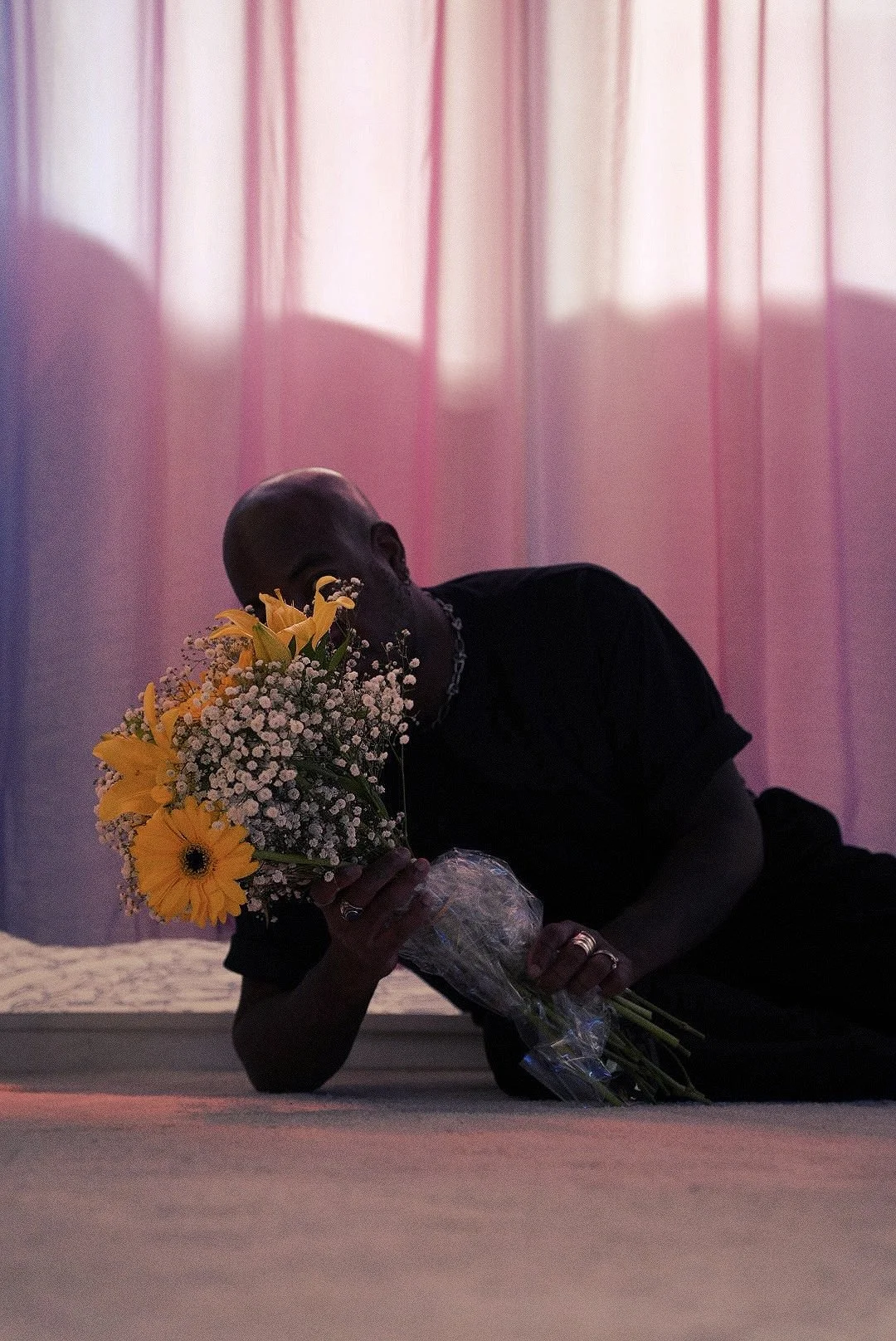Une personne allongée sur le sol, tenant un bouquet de fleurs comprenant des tournesols jaunes et des gypsophiles, devant un rideau rose translucide, avec un éclairage doux. La personne porte des bijoux, y compris une chaîne et des anneaux.