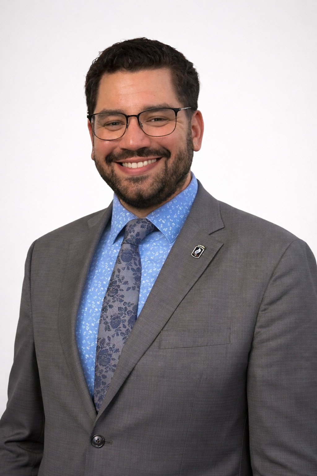A man in a gray suit, blue patterned shirt, and tie, smiling at the camera against a white background.