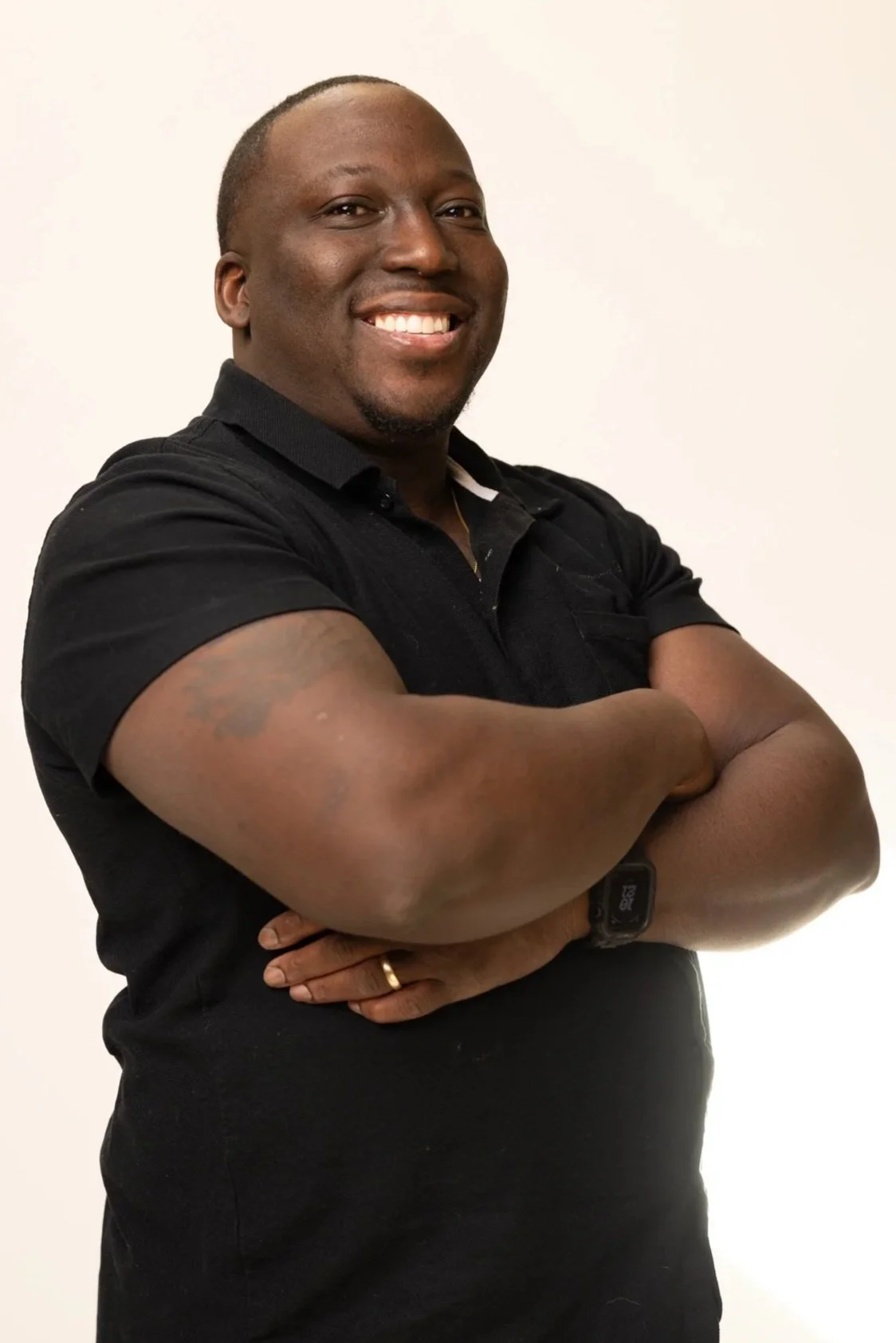 A smiling Black man with a shaved head and goatee wearing a black polo shirt, standing with arms crossed against a neutral background.
