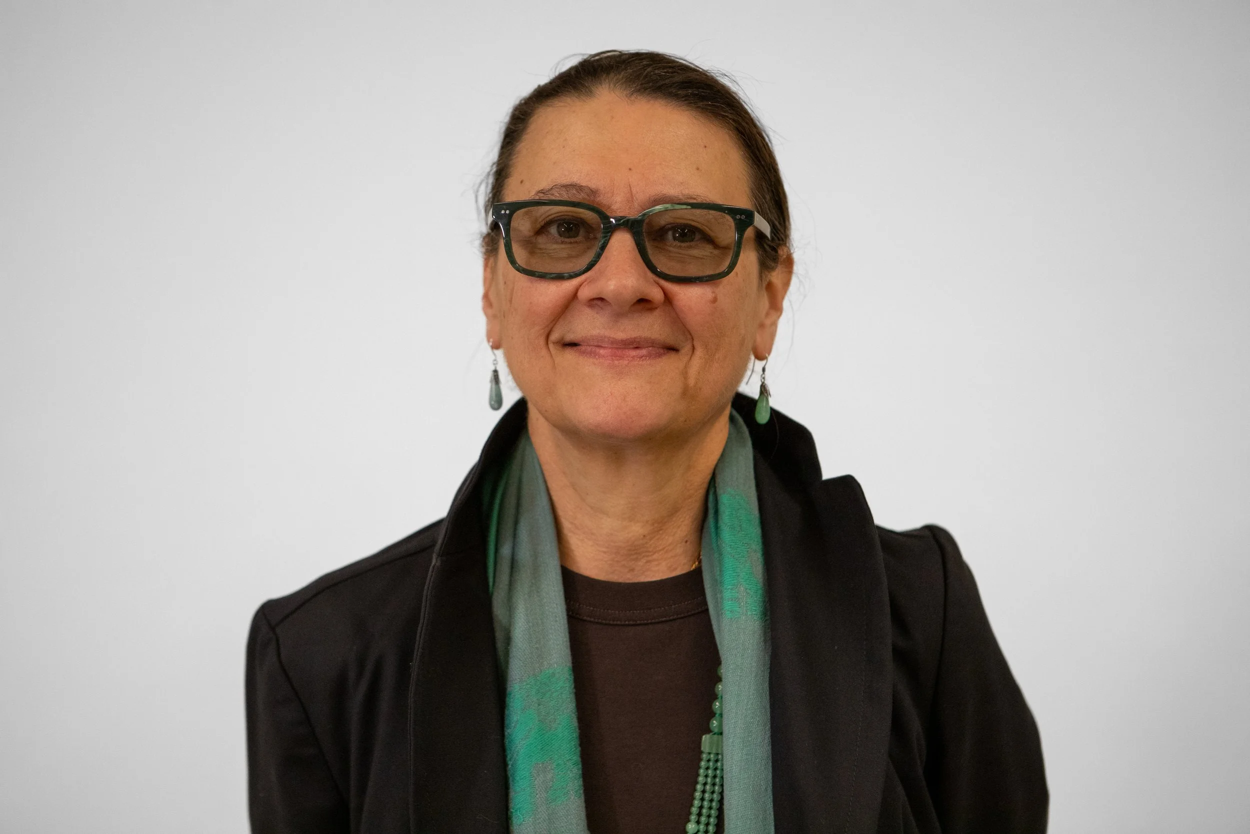 A woman with glasses and earrings, wearing a black blazer and a green scarf, smiling in front of a plain white background.