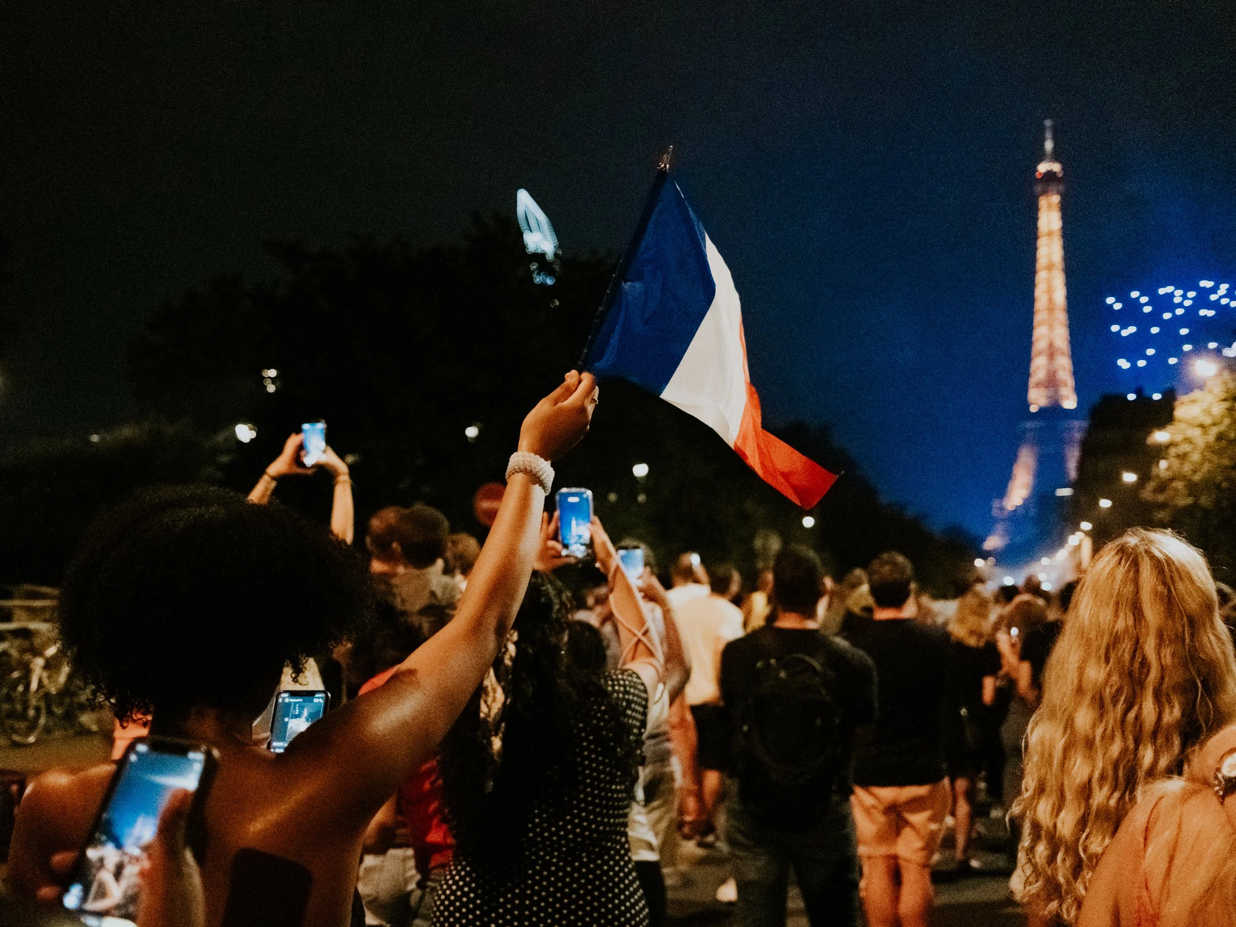 A crowd gathered at night holding up French flags during a celebration, with a large illuminated tower in the background.