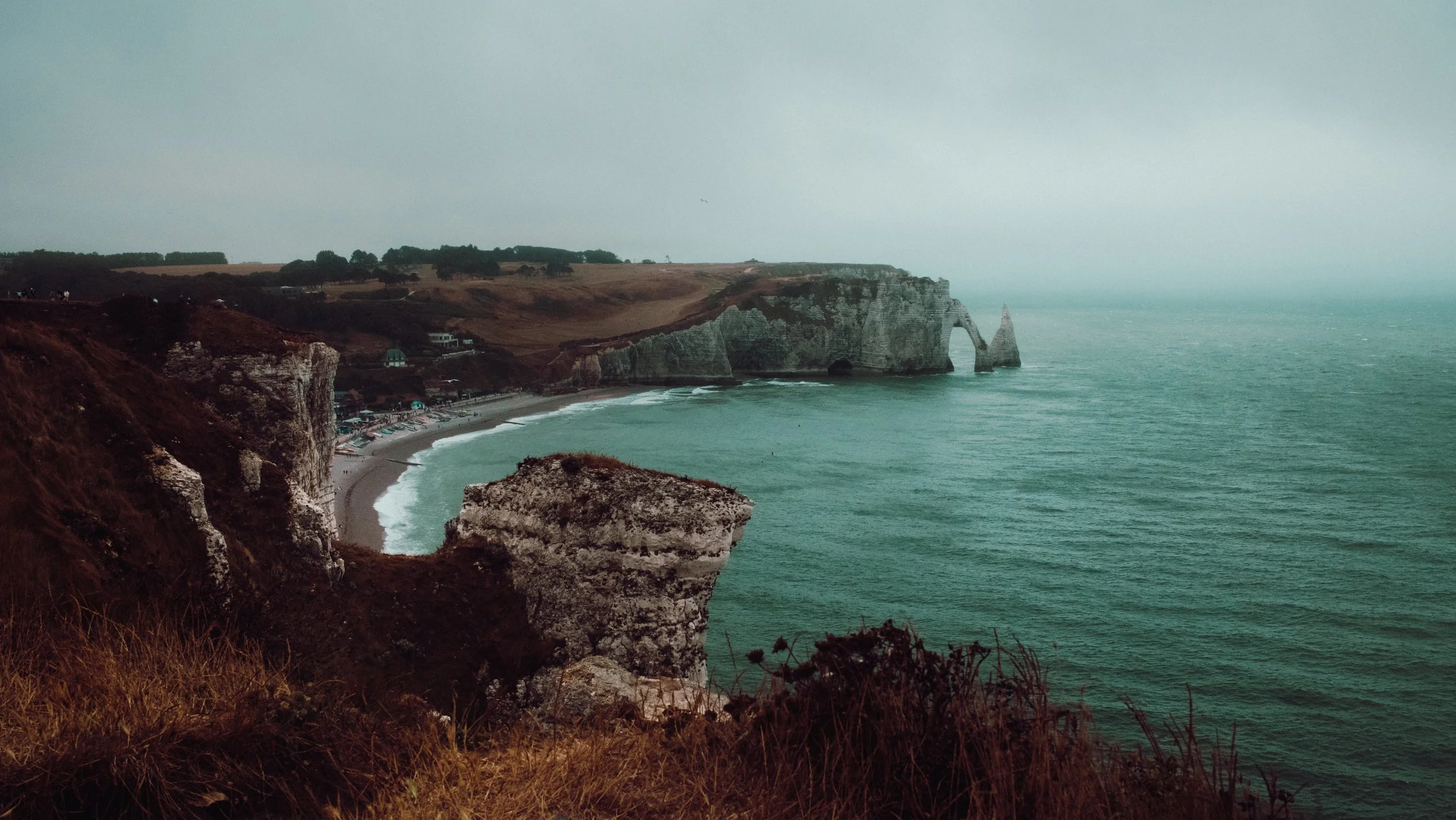 Cliffs and ocean view at a beach with a natural rock arch formation, cloudy sky, and some buildings along the shoreline.