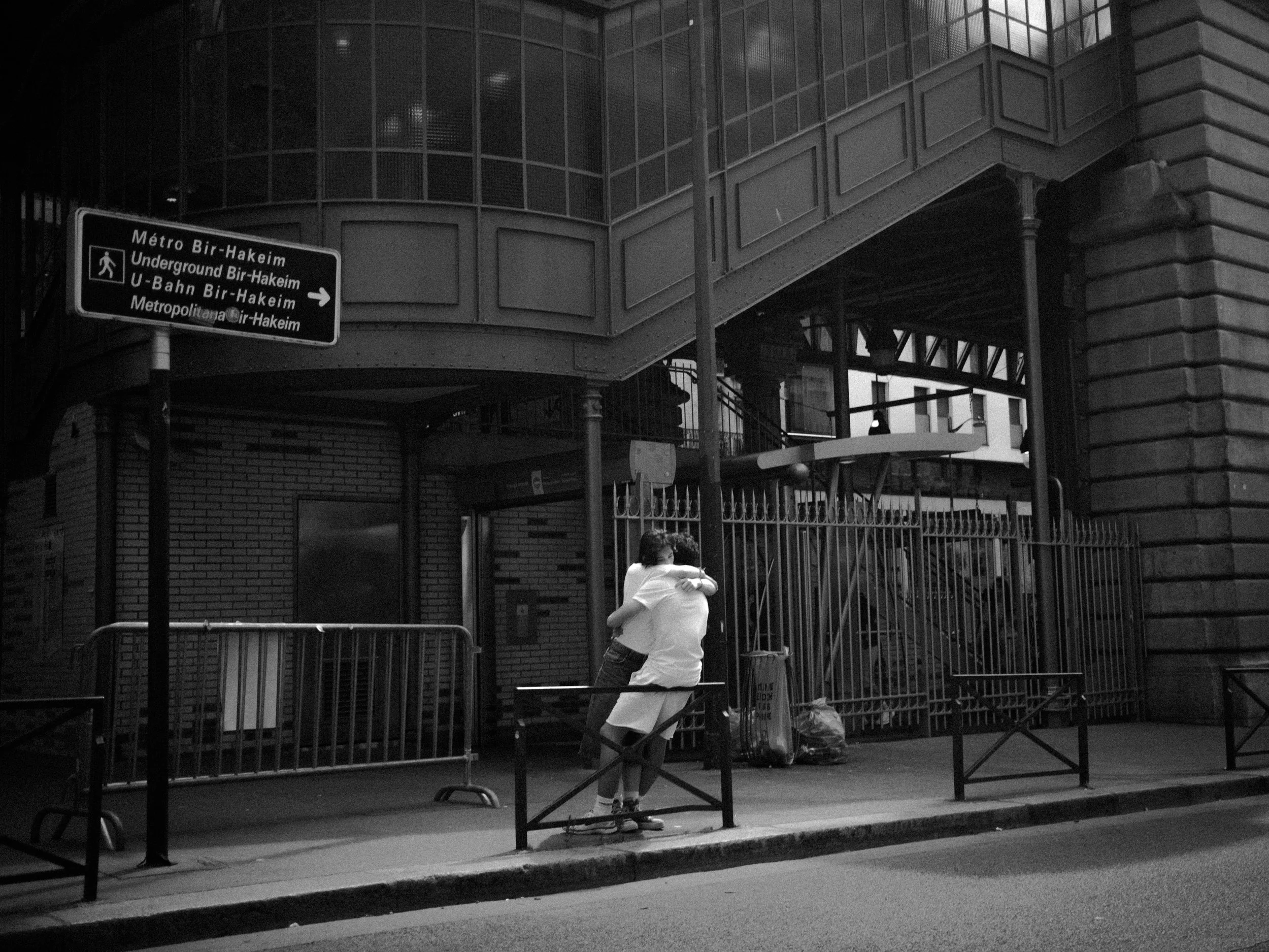Black and white photo of two people hugging in front of a gated area outside a building, with a sign indicating directions to various metro and underground lines. The scene is urban with brick and metal structures.