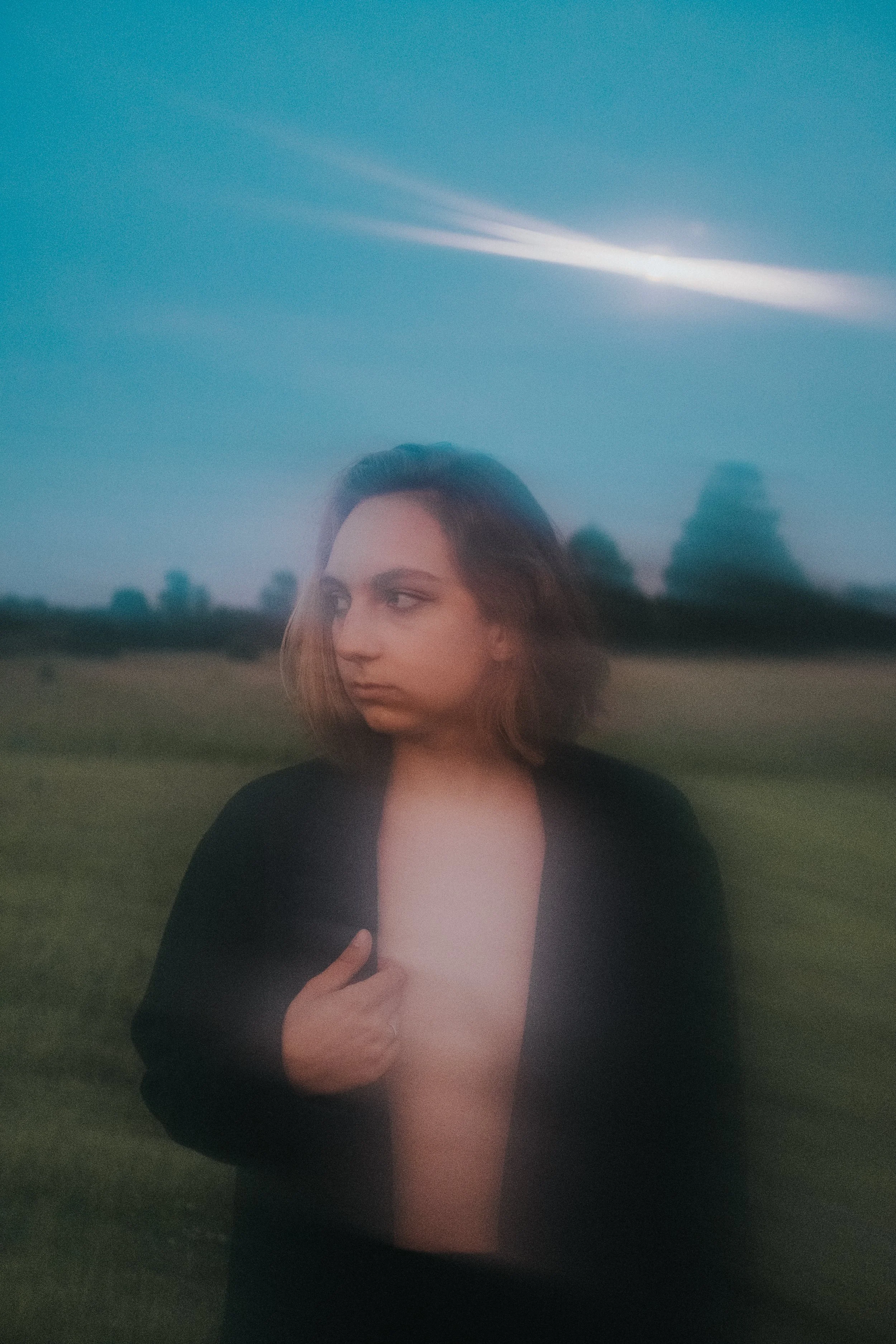 A woman with long hair wearing a black top holds her chest in a grassy field with a blue sky and clouds in the background.