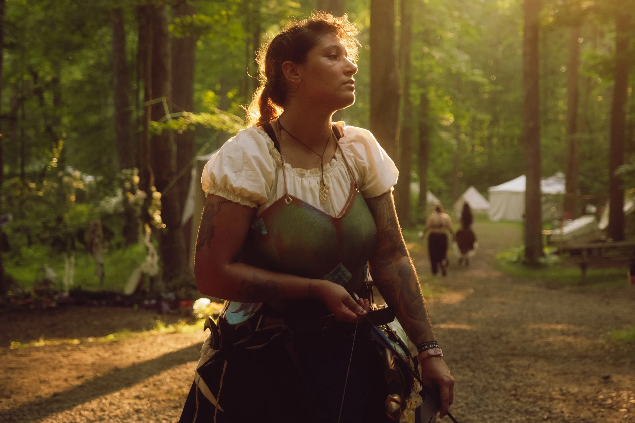 A woman with curly hair, tattoos, and wearing outdoor clothing stands in a forest during sunset, with trees and tents in the background.