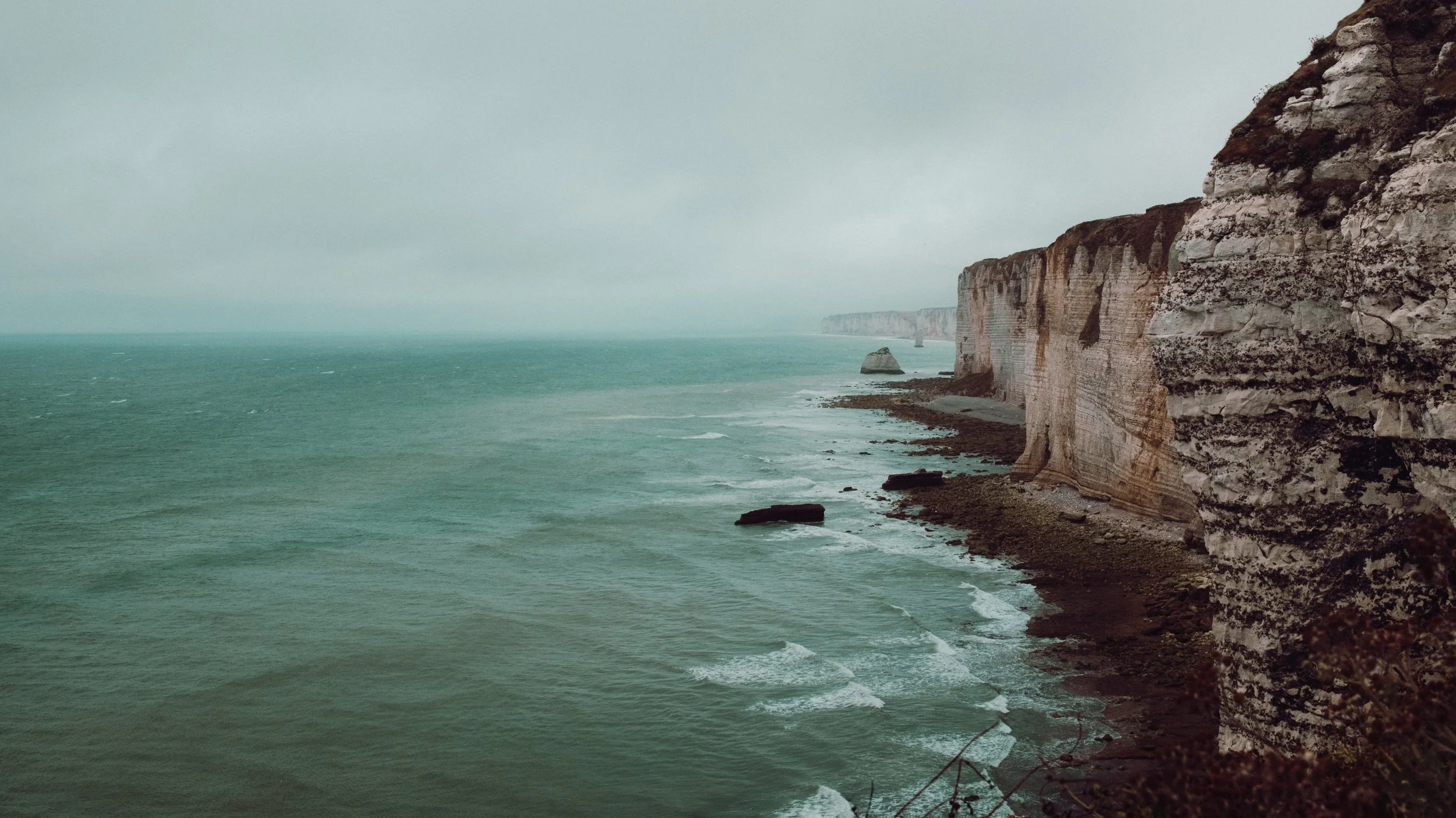 Cliffs along a coastline with rocky formations and greenish ocean waves under overcast sky.