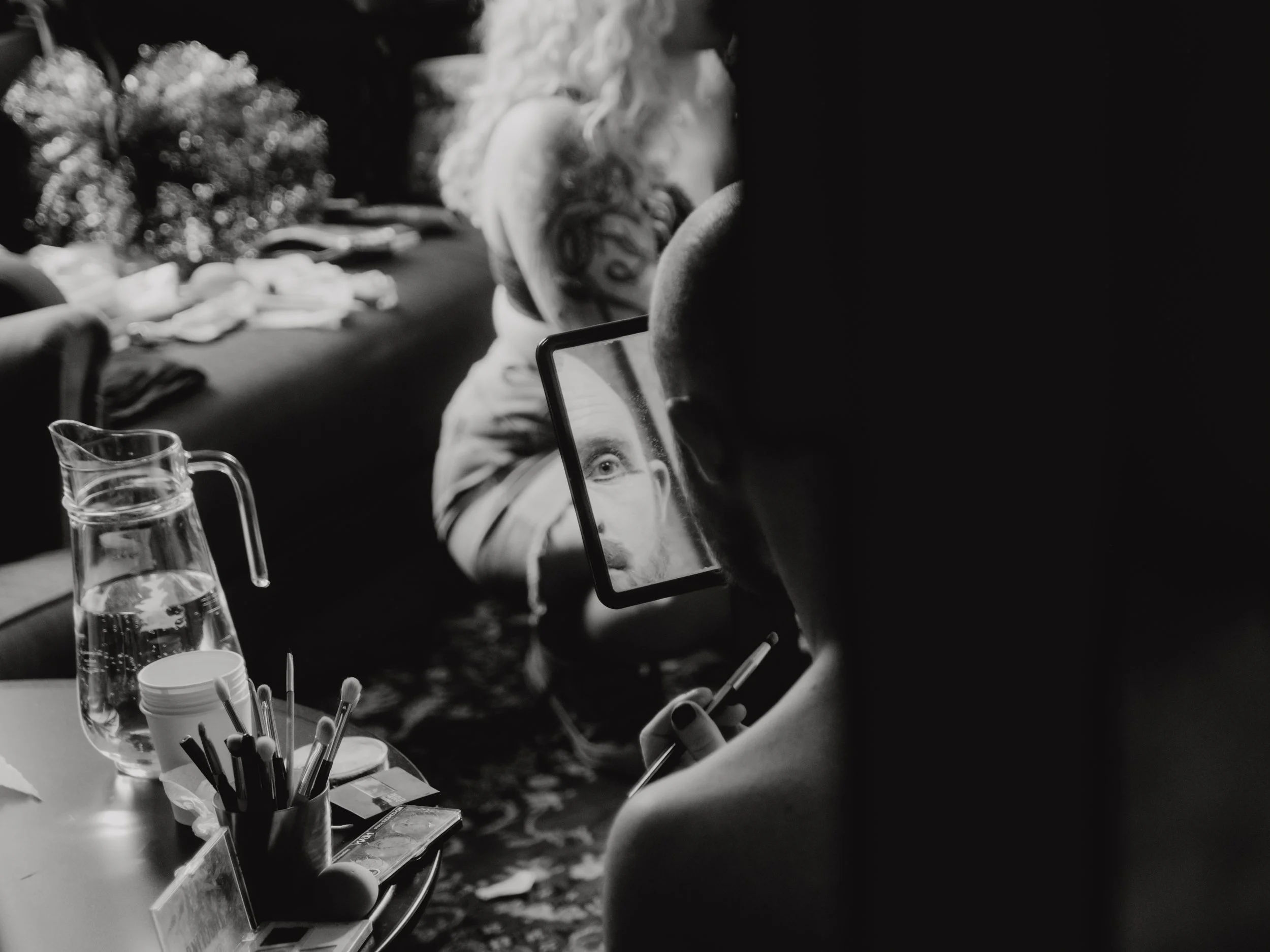 A person with a tattoo on their arm is using their phone on a table with various art supplies, including brushes, a glass of water, and small containers, in a black-and-white photo.