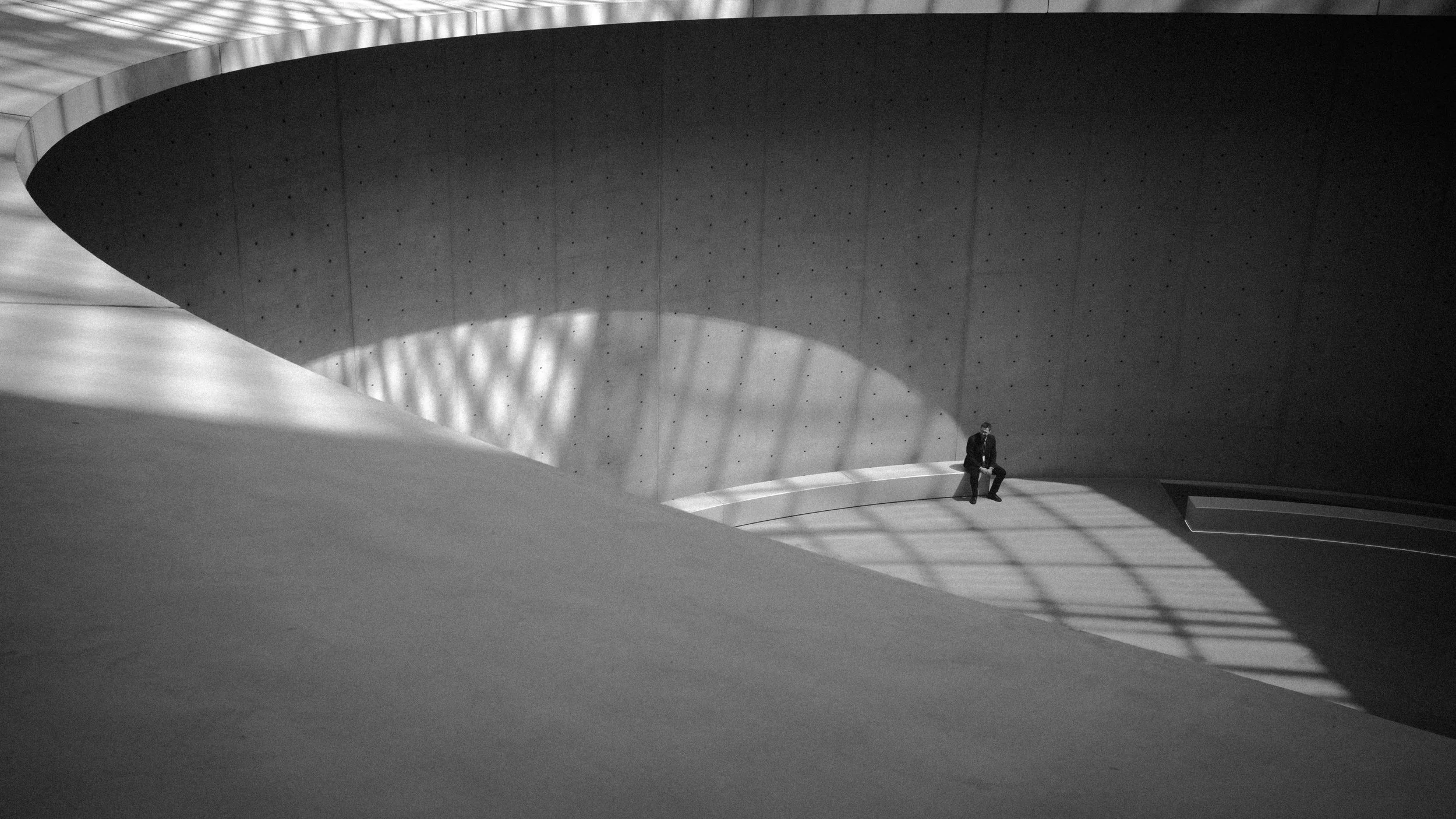 A man in a suit sitting alone on a curved, bench-like structure in a minimalist, modern architectural space with concrete walls and dramatic lighting.