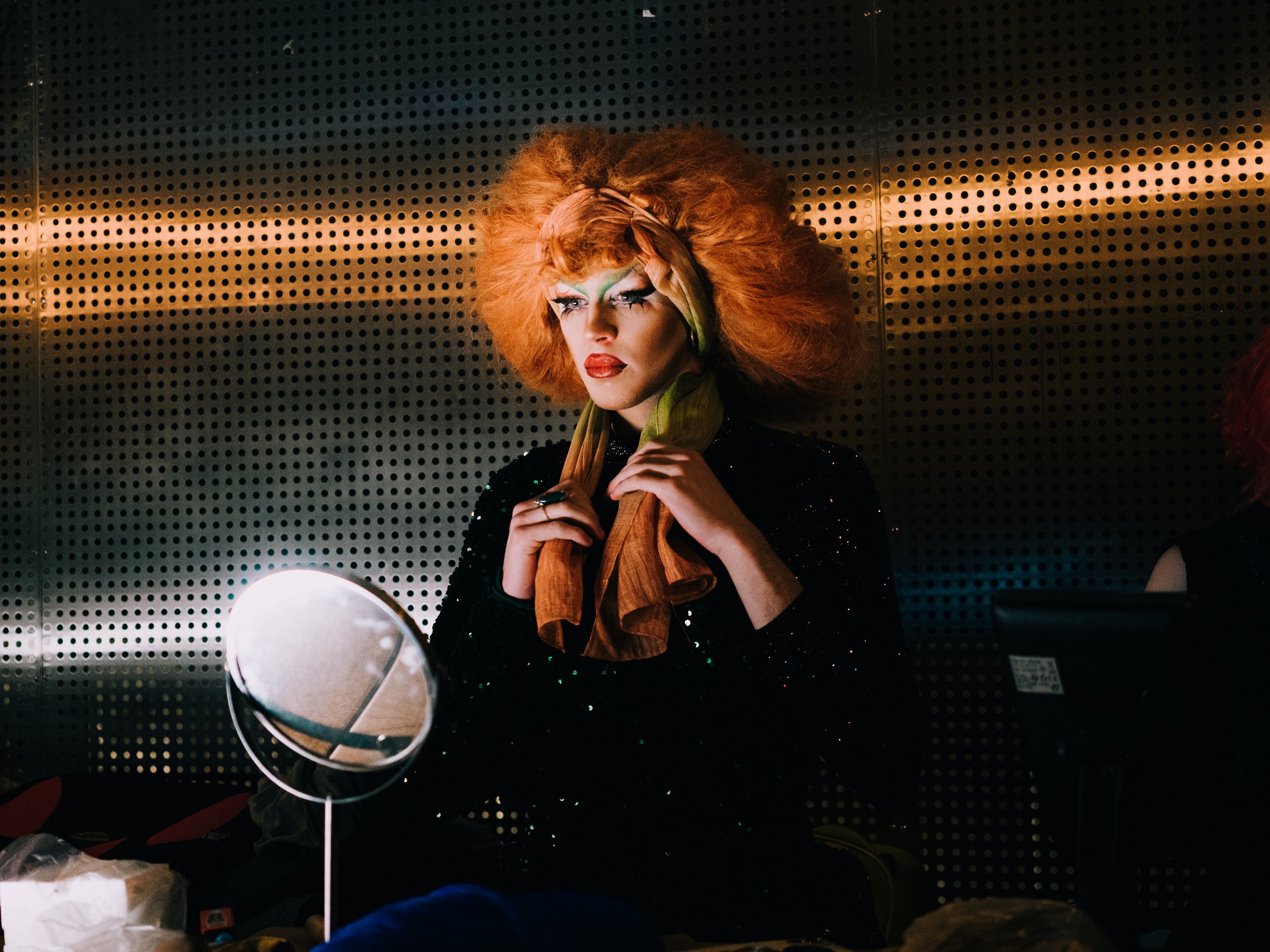 A person with dramatic makeup and large, voluminous orange hair looks at themselves in a mirror. They are dressed in dark, sparkly clothing and are holding a piece of clothing or fabric near their chest. The background features a perforated metal wal