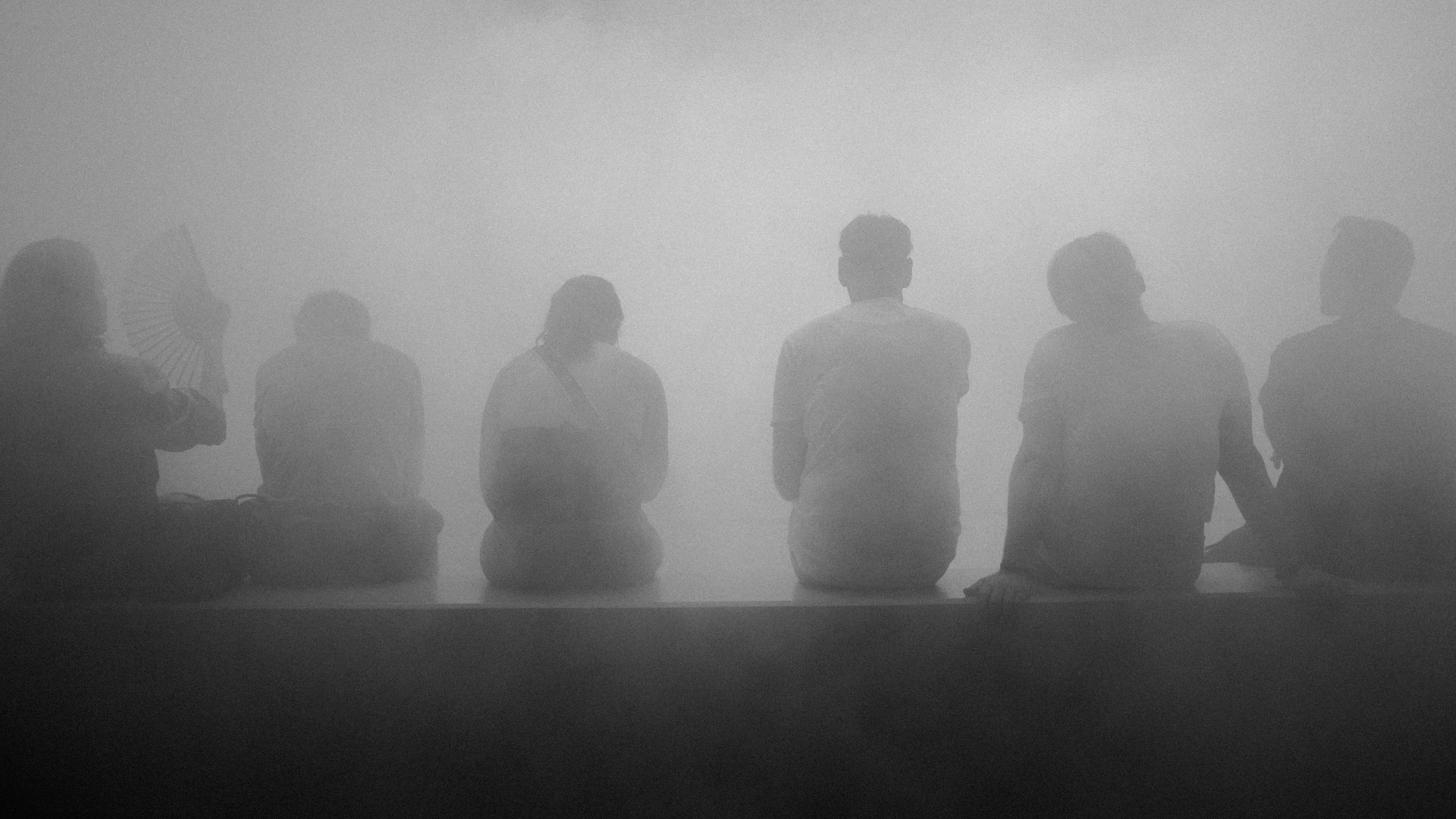 Silhouettes of seven people sitting, some holding umbrellas, facing a foggy or smoky background.